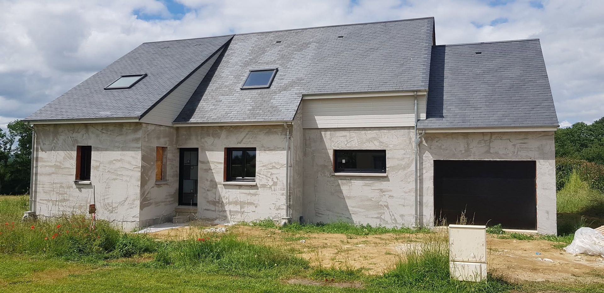 Maison partiellement construite avec toit gris, façade en pierre inachevée et garage ouvert sur un terrain gazonné.