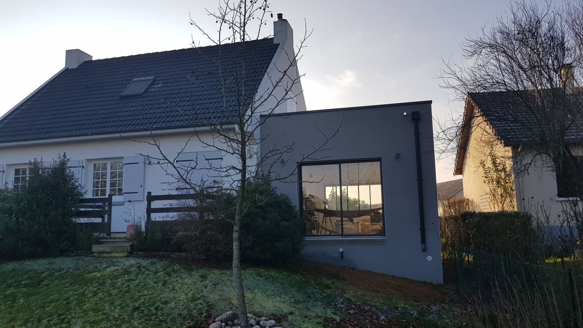 Maison au crépuscule avec une extension moderne éclairée, un jardin et des arbres dénudés au premier plan.