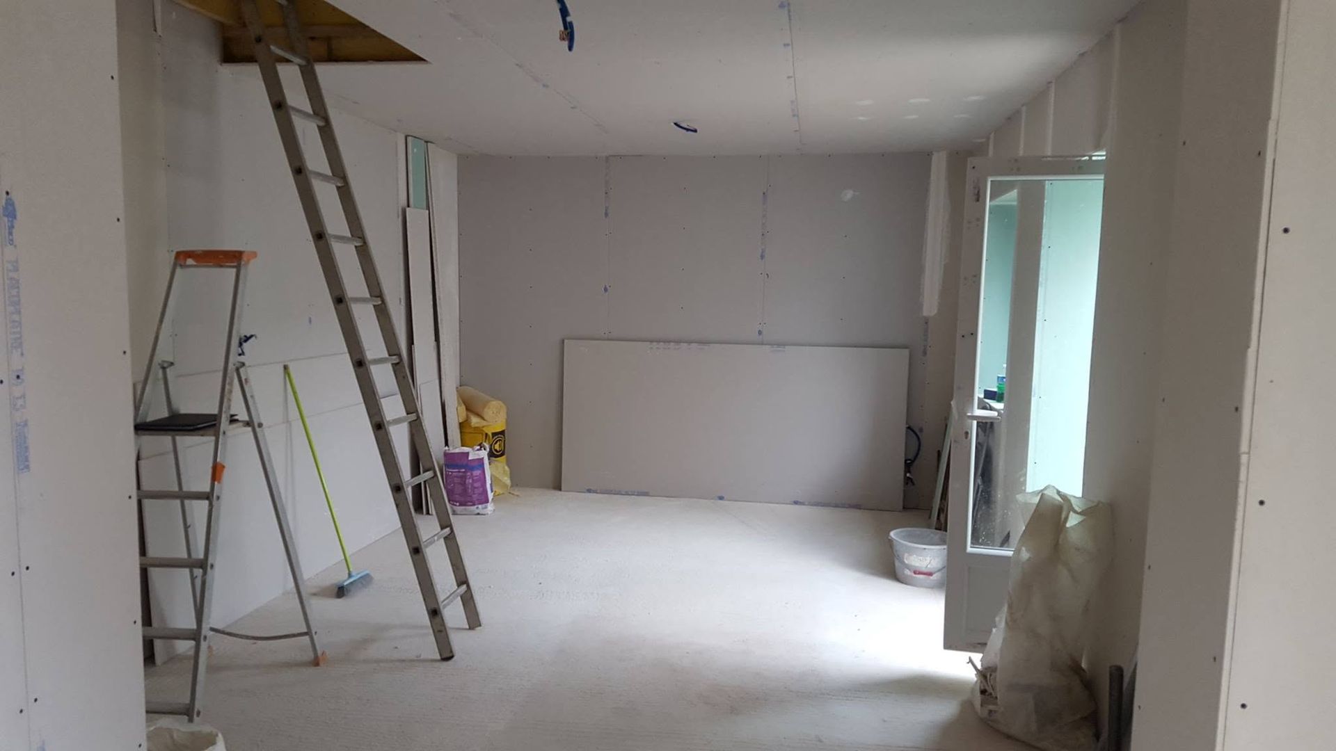 Chantier de rénovation avec échelles, cloisons sèches et murs non finis dans une pièce lumineuse et vide.