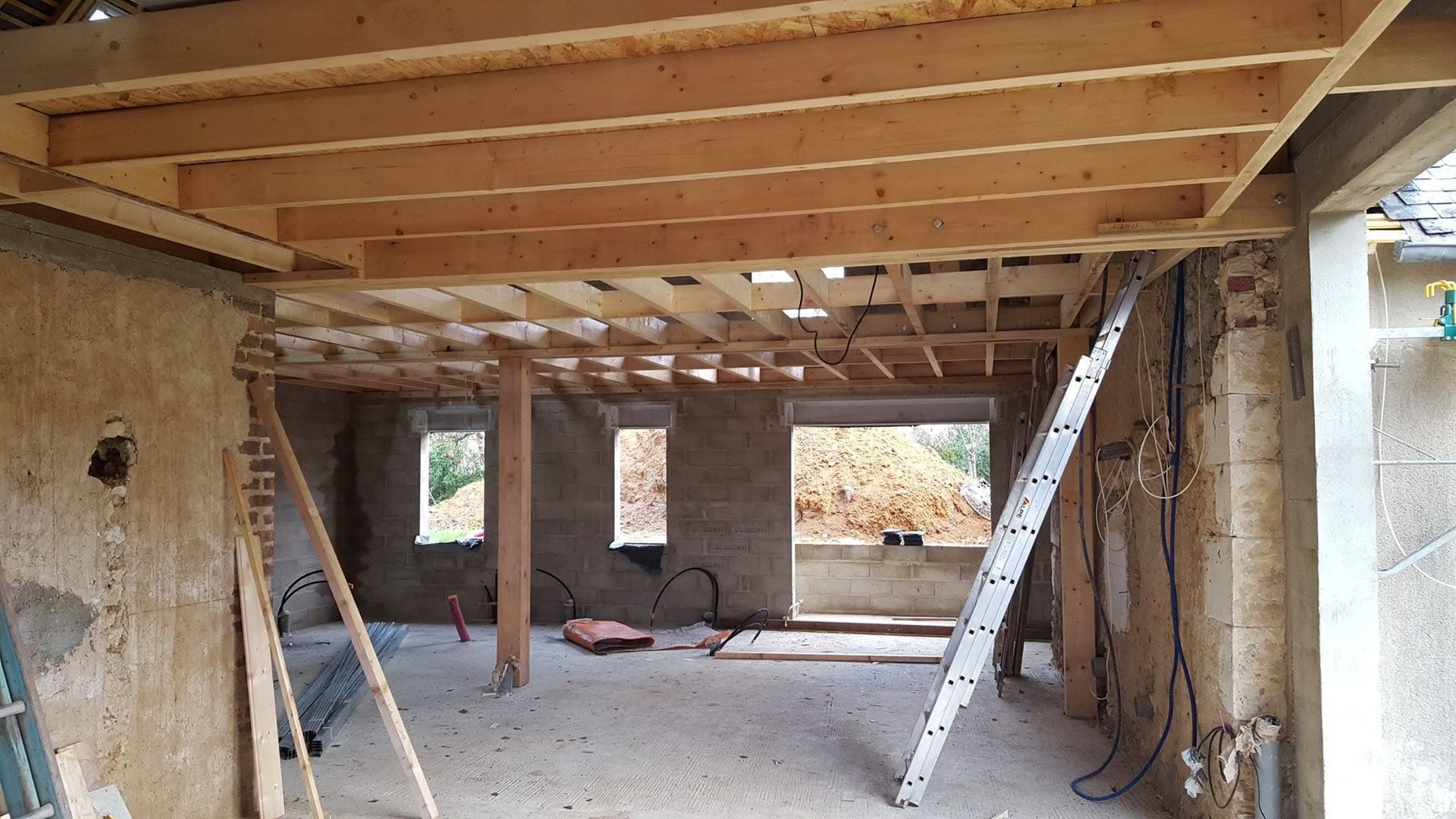 Pièce partiellement construite avec poutres de plafond en bois apparentes, murs en pierre et deux échelles sur un sol en béton.