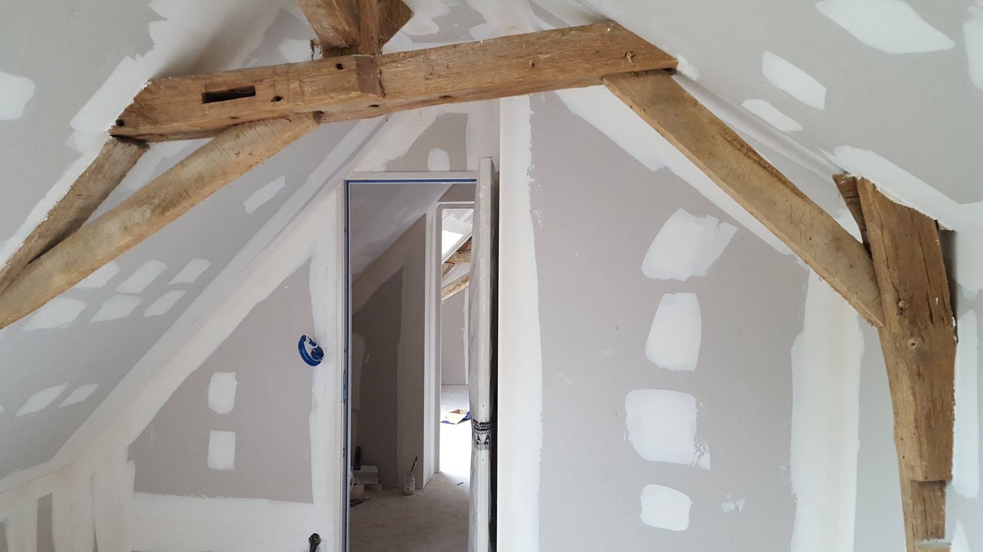 Pièce mansardée avec poutres en bois apparentes et plaques de plâtre blanches autour d'une porte.
