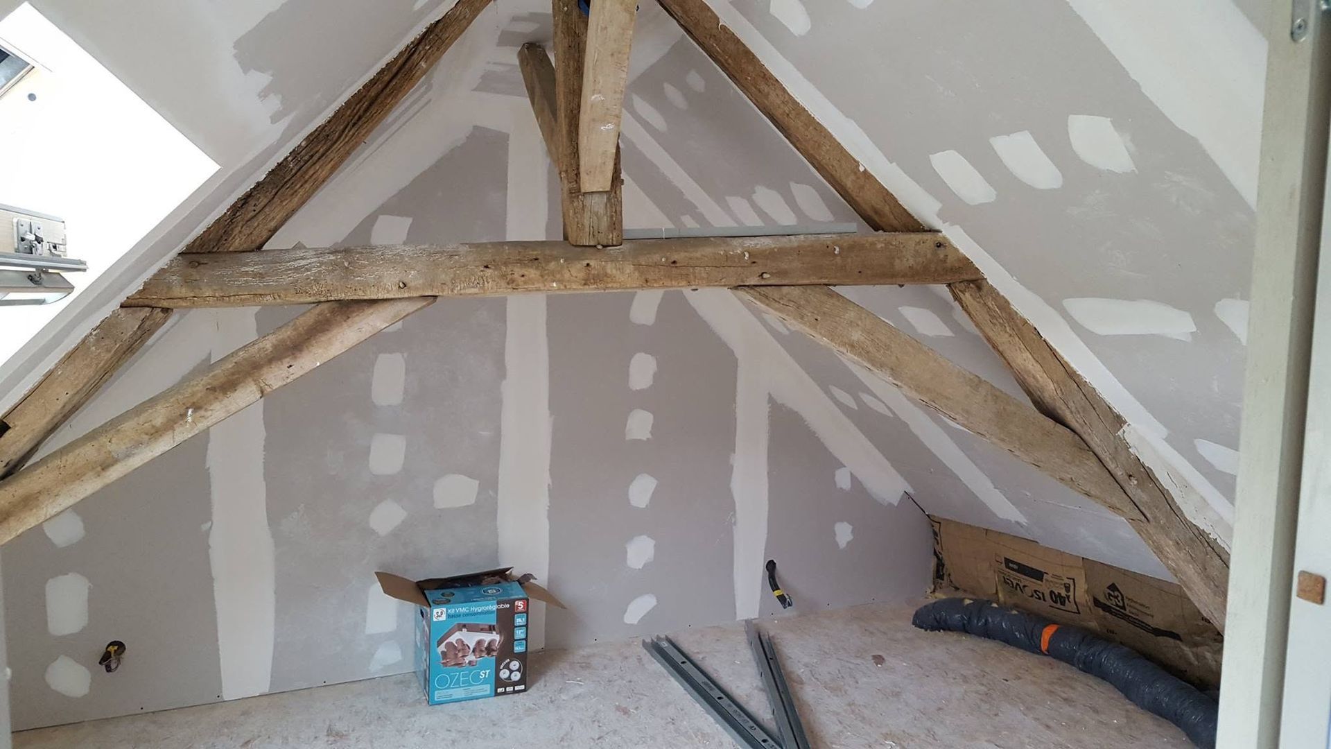 Grenier non aménagé avec poutres en bois apparentes, cloisons sèches rapiécées et matériaux de construction au sol.