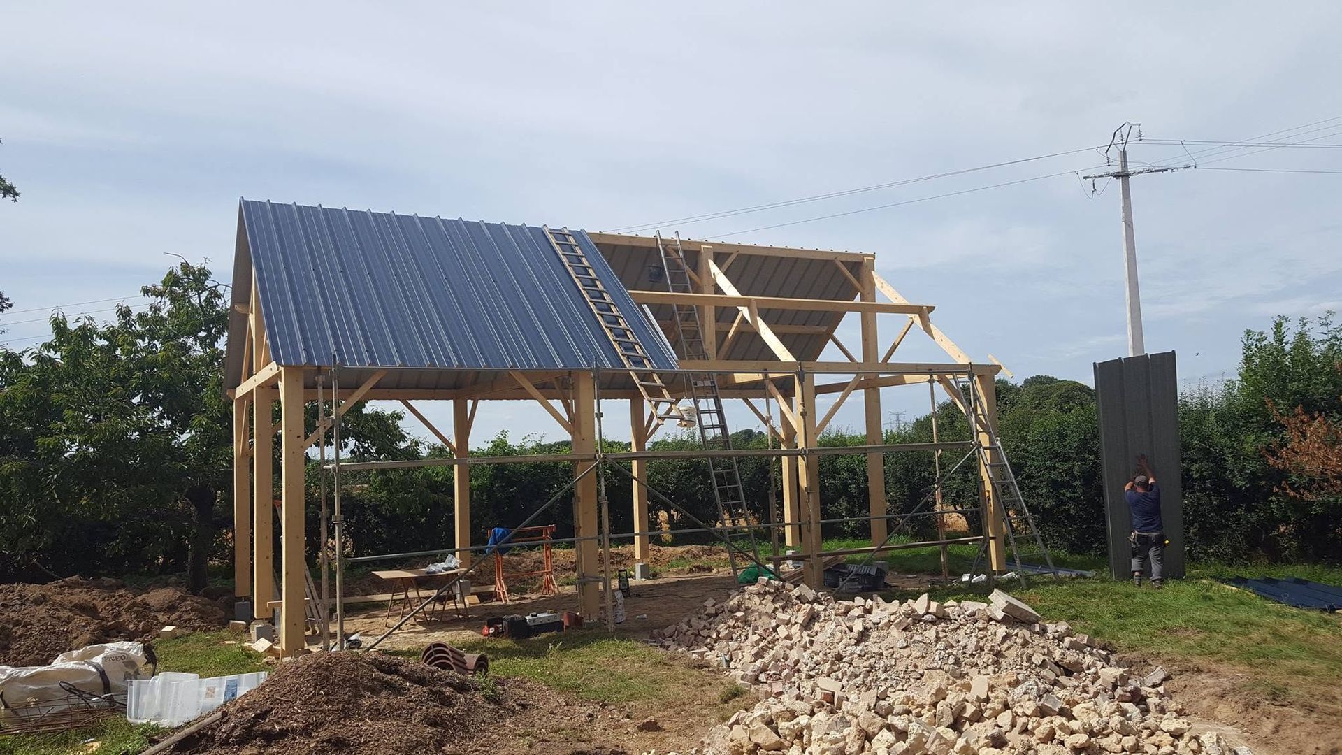 Maison à ossature bois partiellement construite, avec un toit en métal sombre, dans un champ, des tas de bois et de débris à proximité.