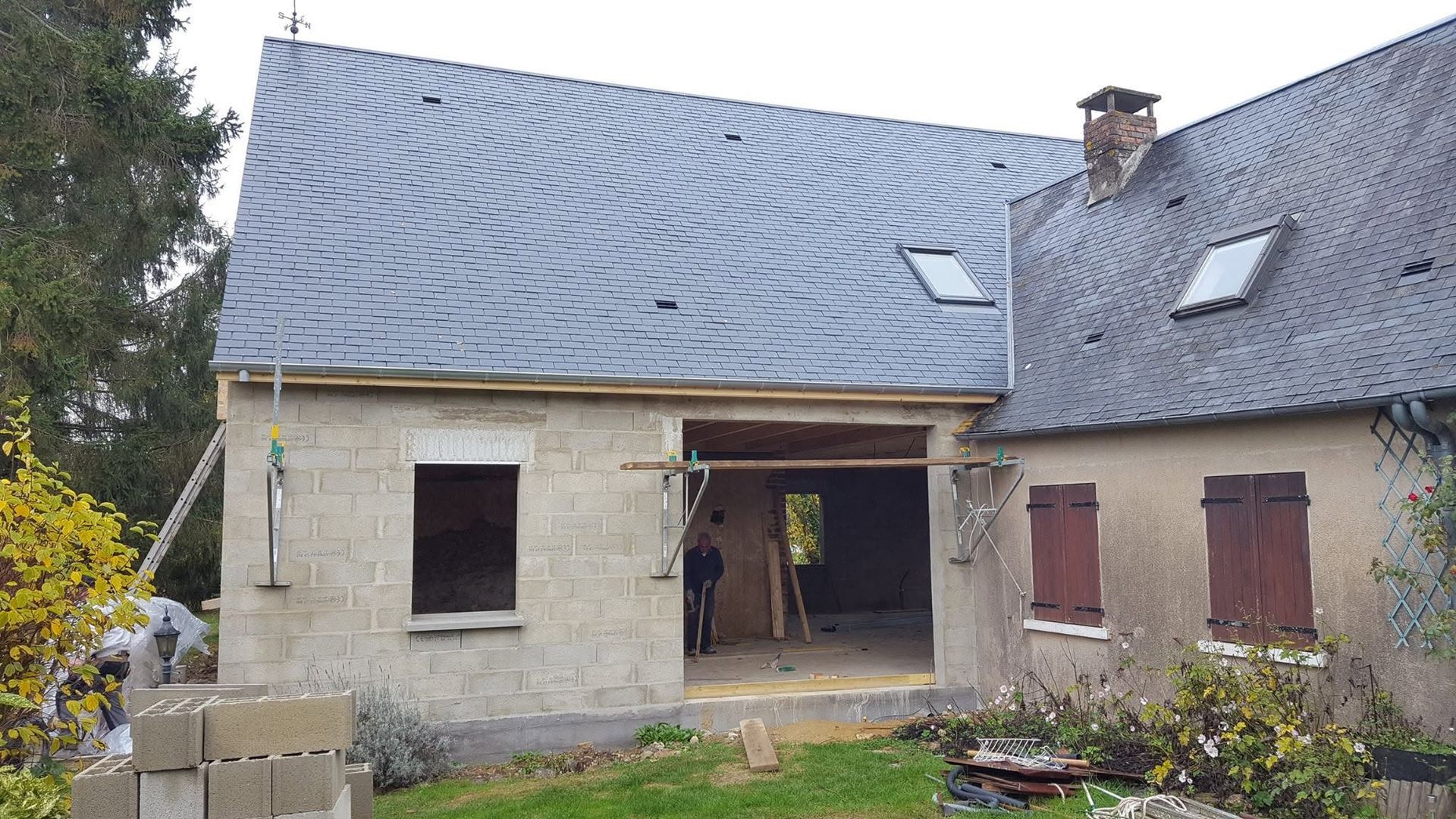 Maison en pierre partiellement rénovée, avec un toit gris, une porte centrale ouverte et une cour envahie par la végétation.