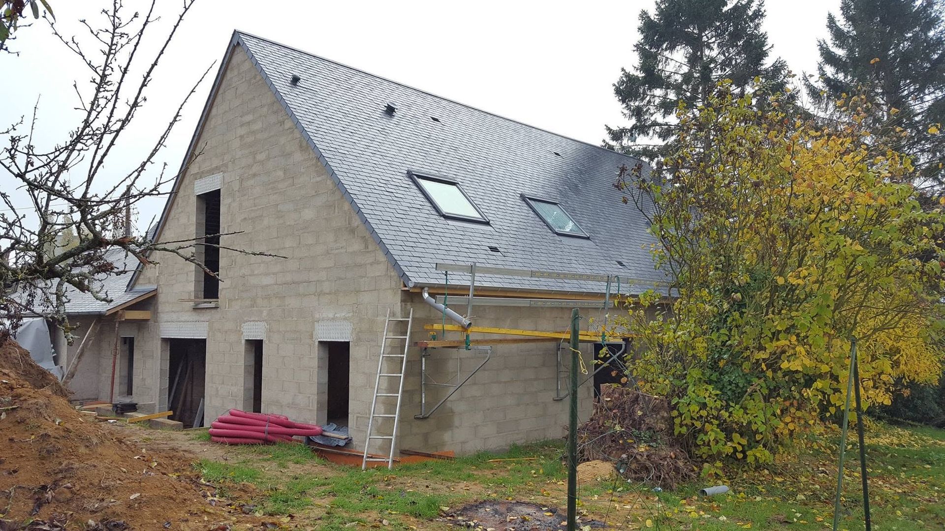 Maison en pierre avec un toit métallique pentu et des échafaudages dans une cour verdoyante en automne