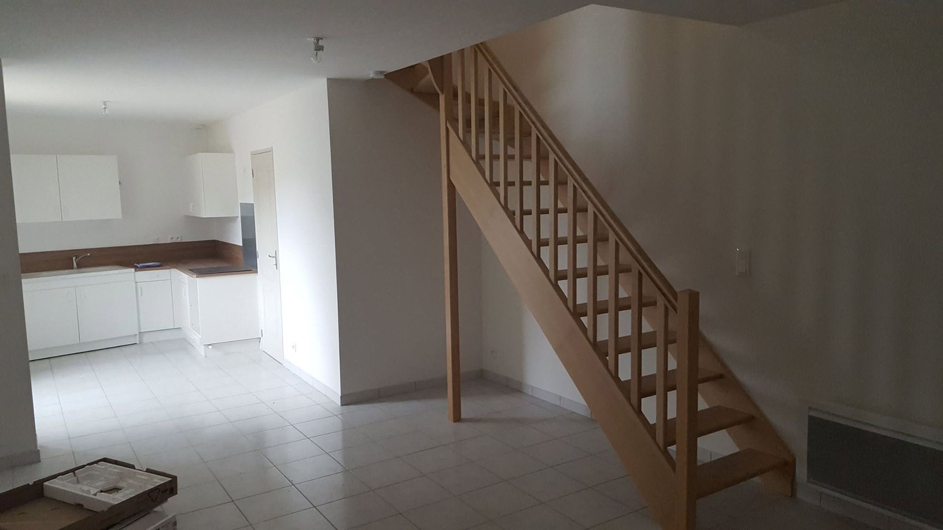 Pièce vide avec sol carrelé, escalier en bois et coin cuisine ouvert en arrière-plan.