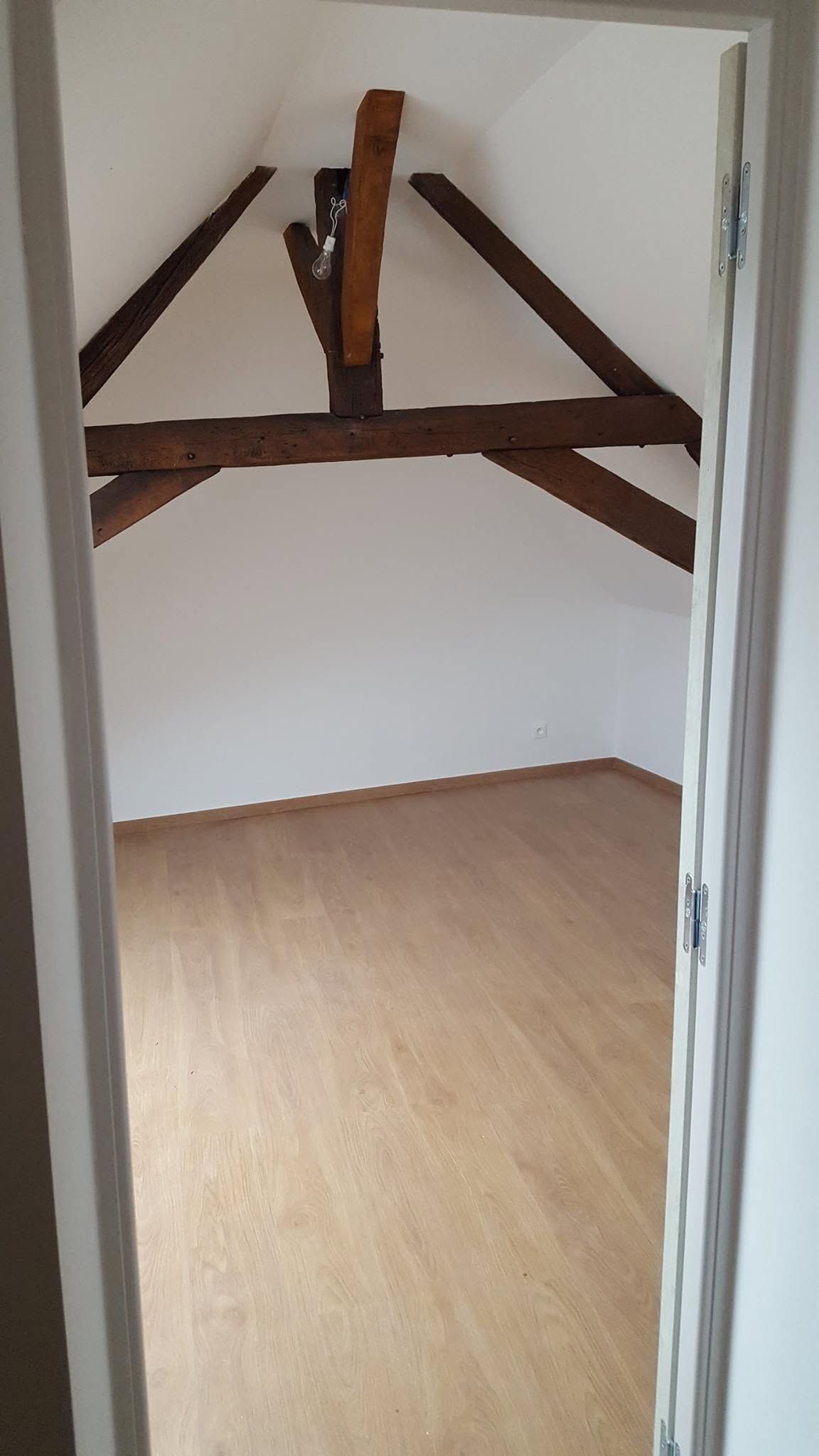 Petite pièce vide avec parquet, murs blancs et poutres apparentes sombres dans l'encadrement de la porte.