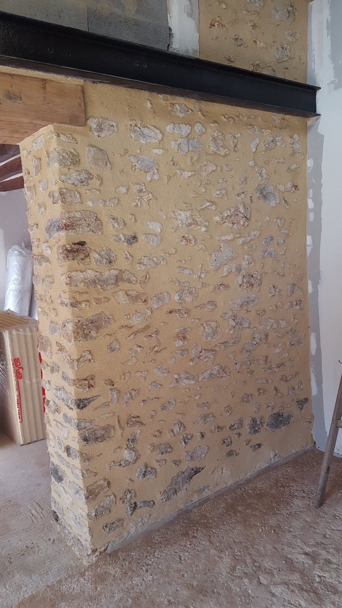 Gros plan sur un coin de mur beige texturé avec un rebord noir et des débris de construction au sol.