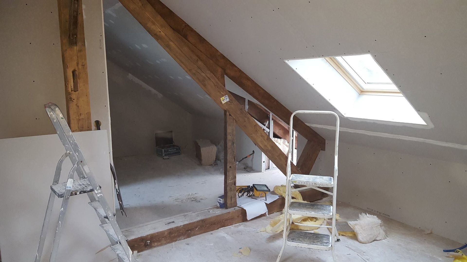 Grenier en rénovation avec poutres apparentes, échelle et fenêtre de toit.