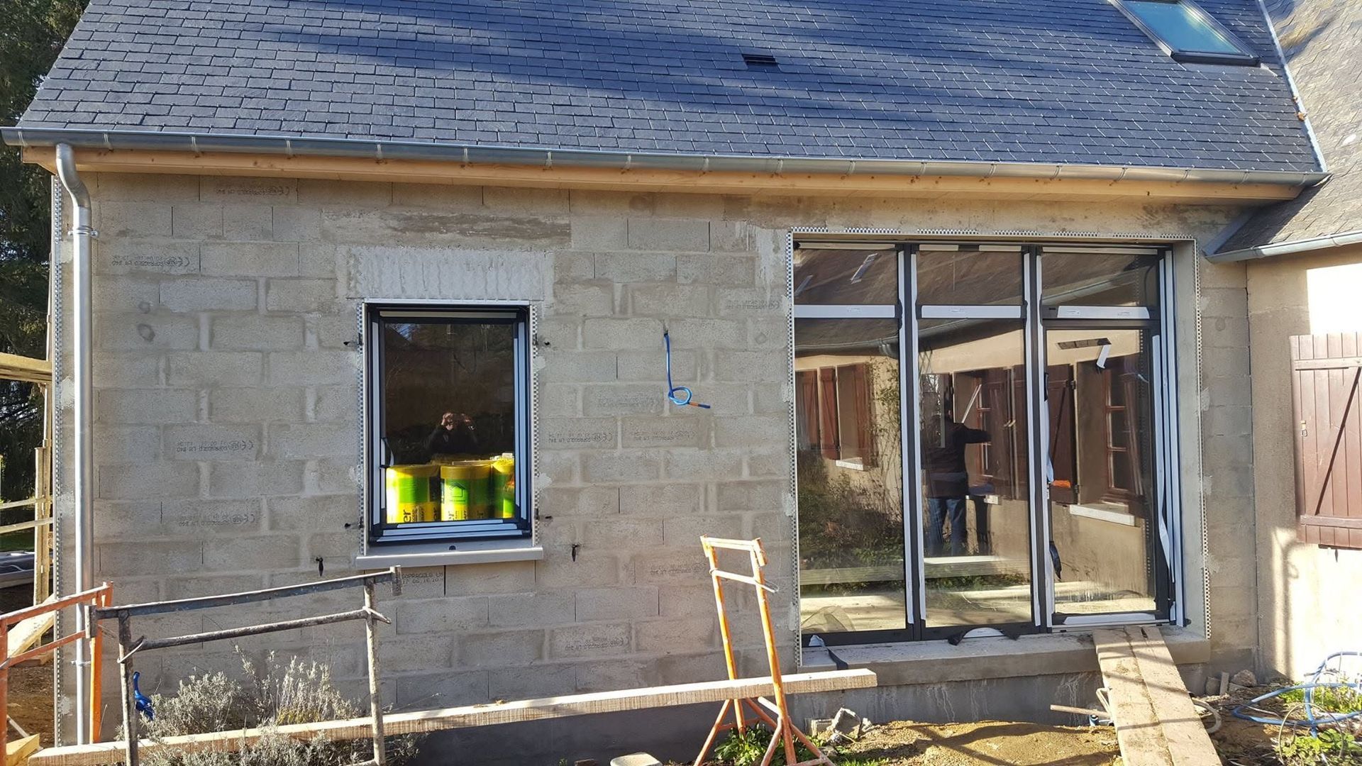 Maison en pierre en construction, façade en pierre avec toit bleu, murs inachevés, fenêtres et portes-fenêtres ouvertes, matériaux de construction à l'extérieur.