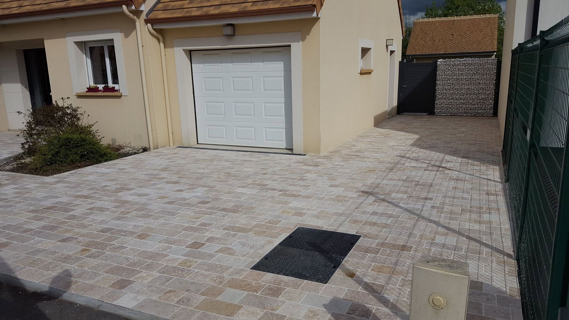 Allée résidentielle pavée de pierres claires, porte de garage blanche et portail vert à côté d'une maison