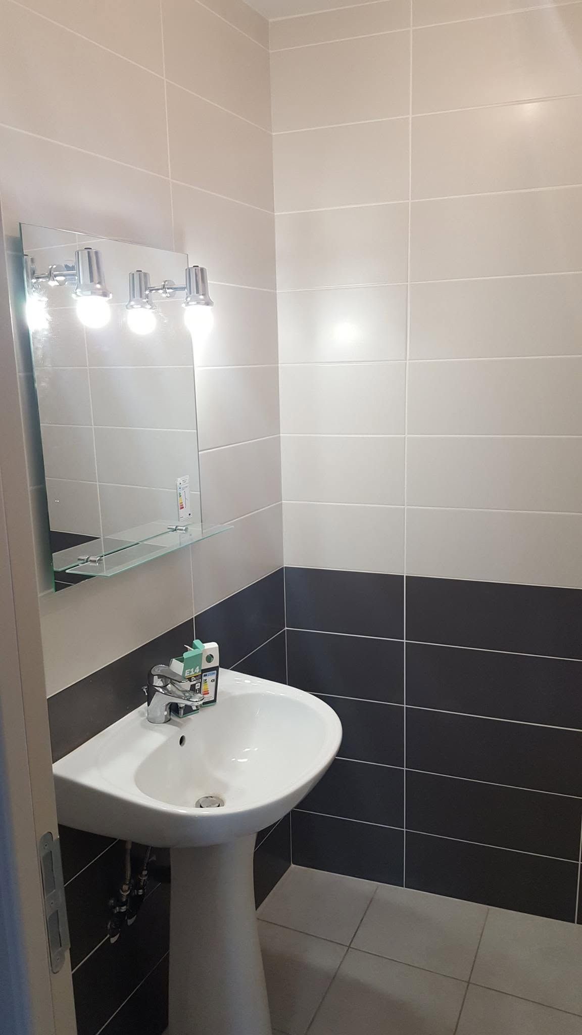 Petite salle de bains carrelée avec miroir mural, éclairage et lavabo blanc avec distributeur de savon.