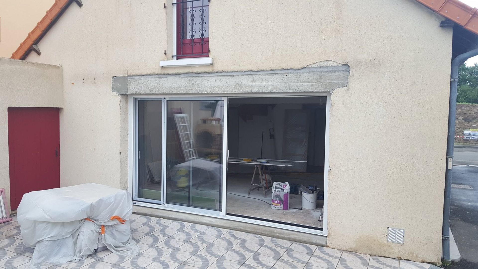 Extérieur de la maison avec de grandes portes coulissantes en verre, une porte latérale rouge et des matériaux de construction dans la cour.
