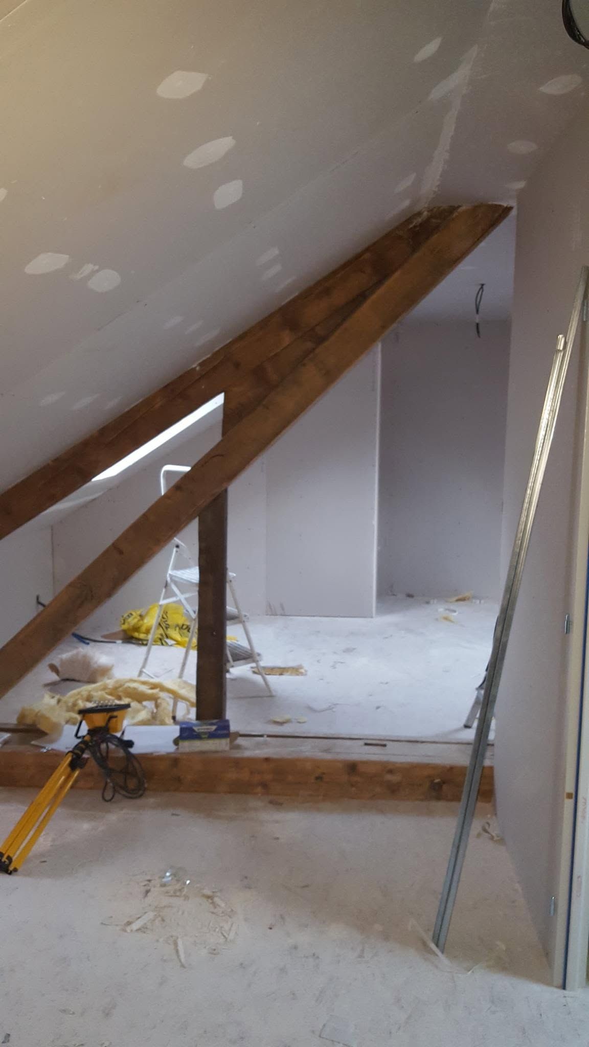 Grenier en rénovation avec poutres apparentes, plaques de plâtre réparées et outils de chantier au sol.