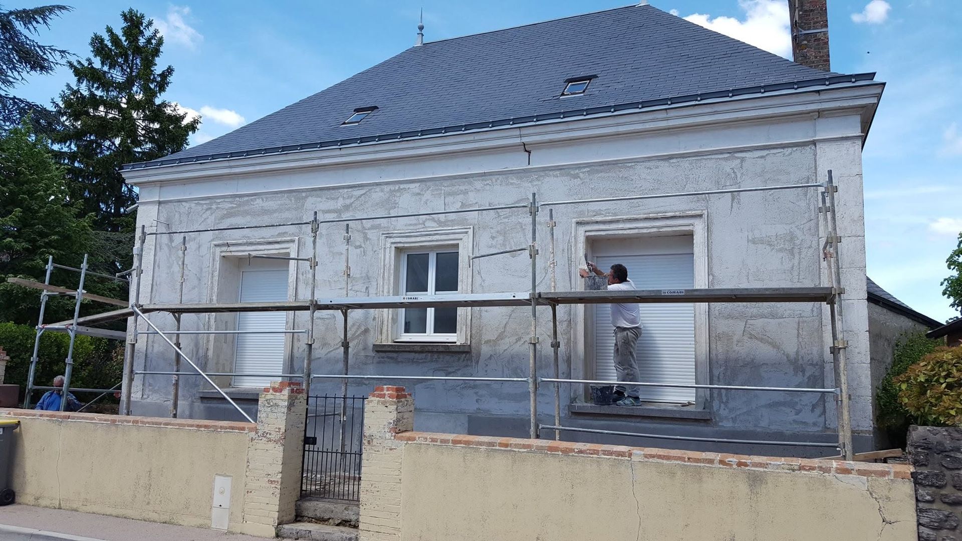 Un ouvrier peint la façade d'une maison blanche derrière des échafaudages et une clôture.