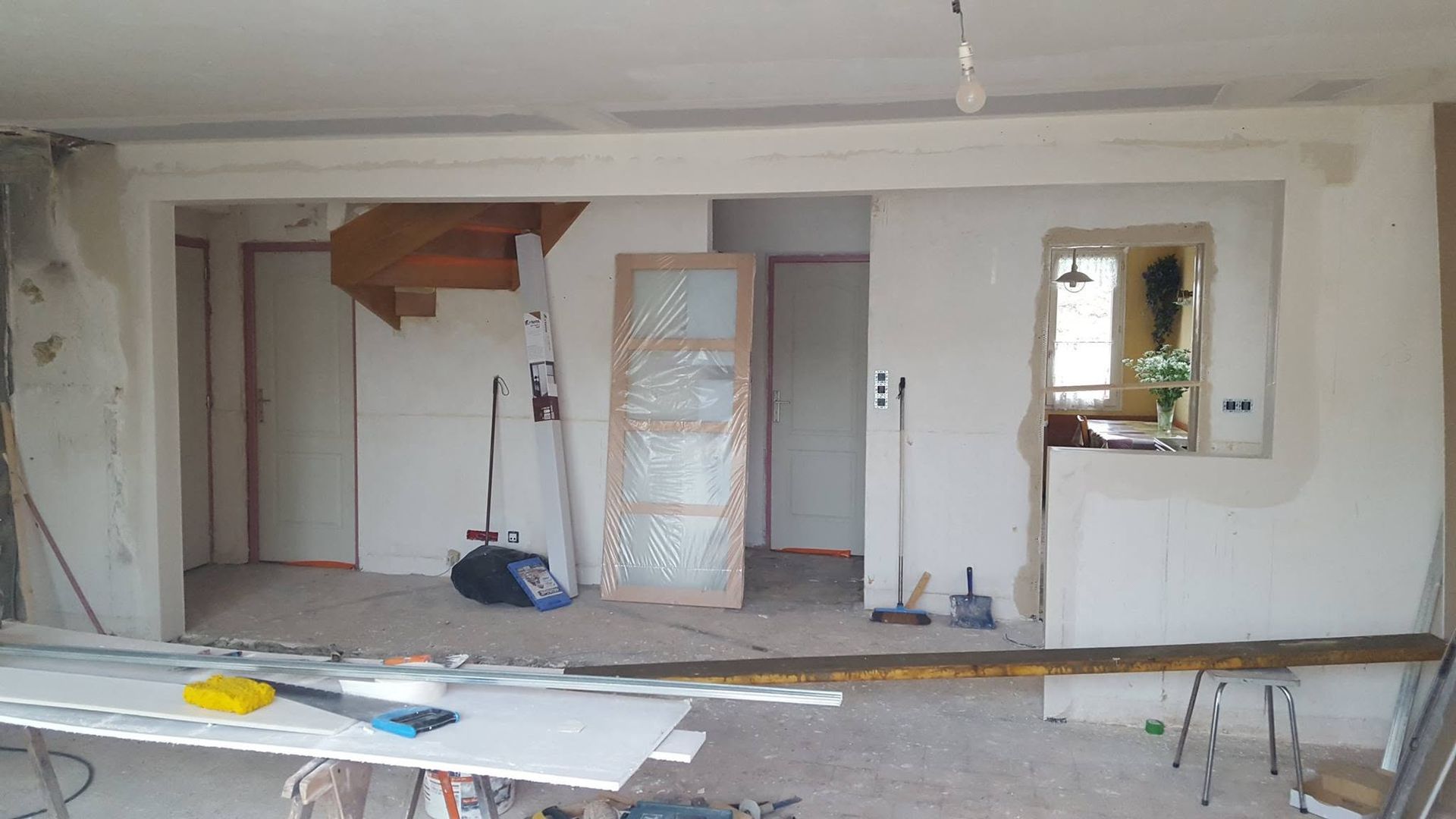 Pièce rénovée avec murs blancs, poutres apparentes, porte ouverte, fenêtre et matériaux de construction au sol.