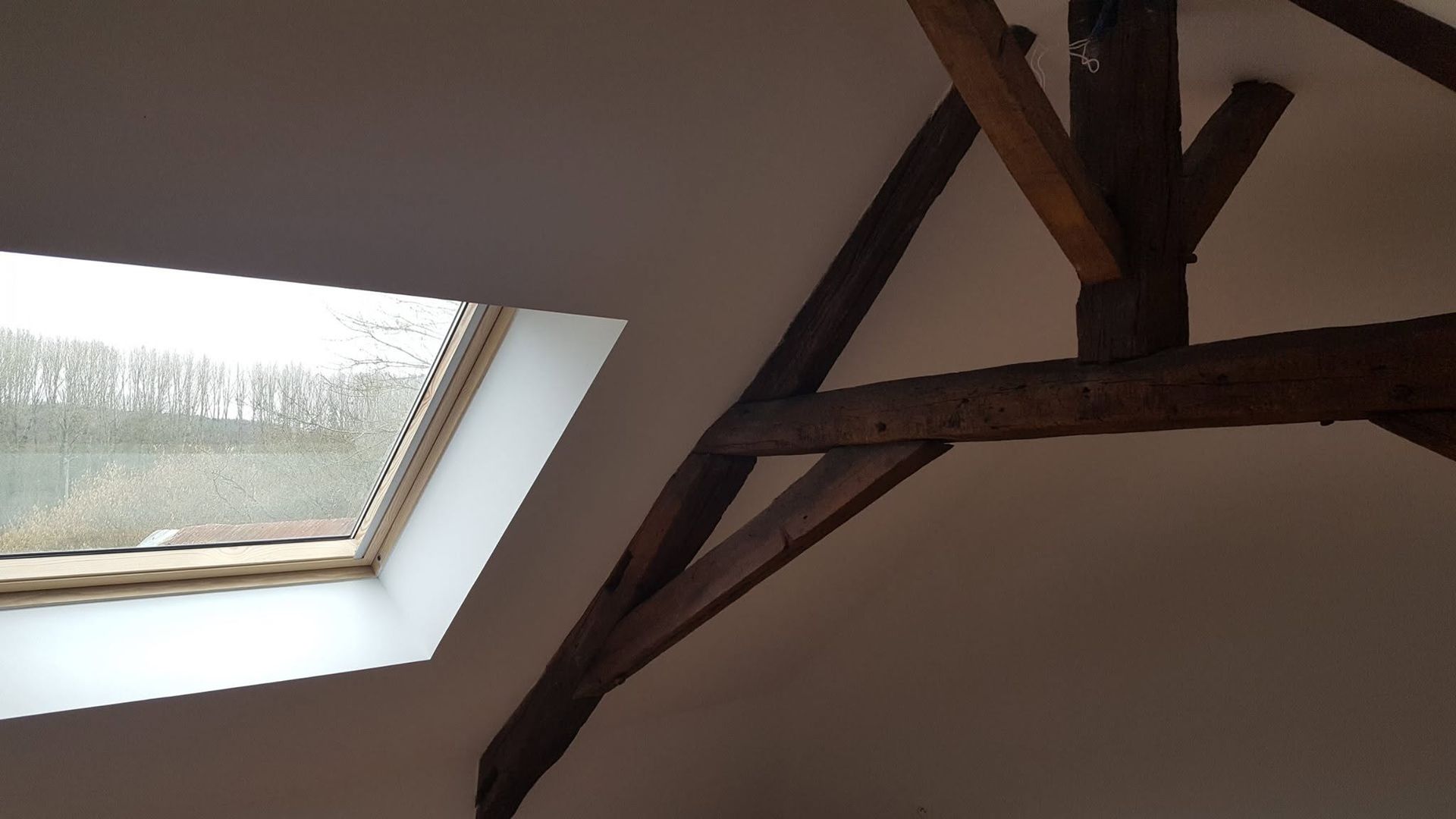 Puits de lumière incliné à côté des poutres apparentes en bois sombre du plafond dans une pièce.