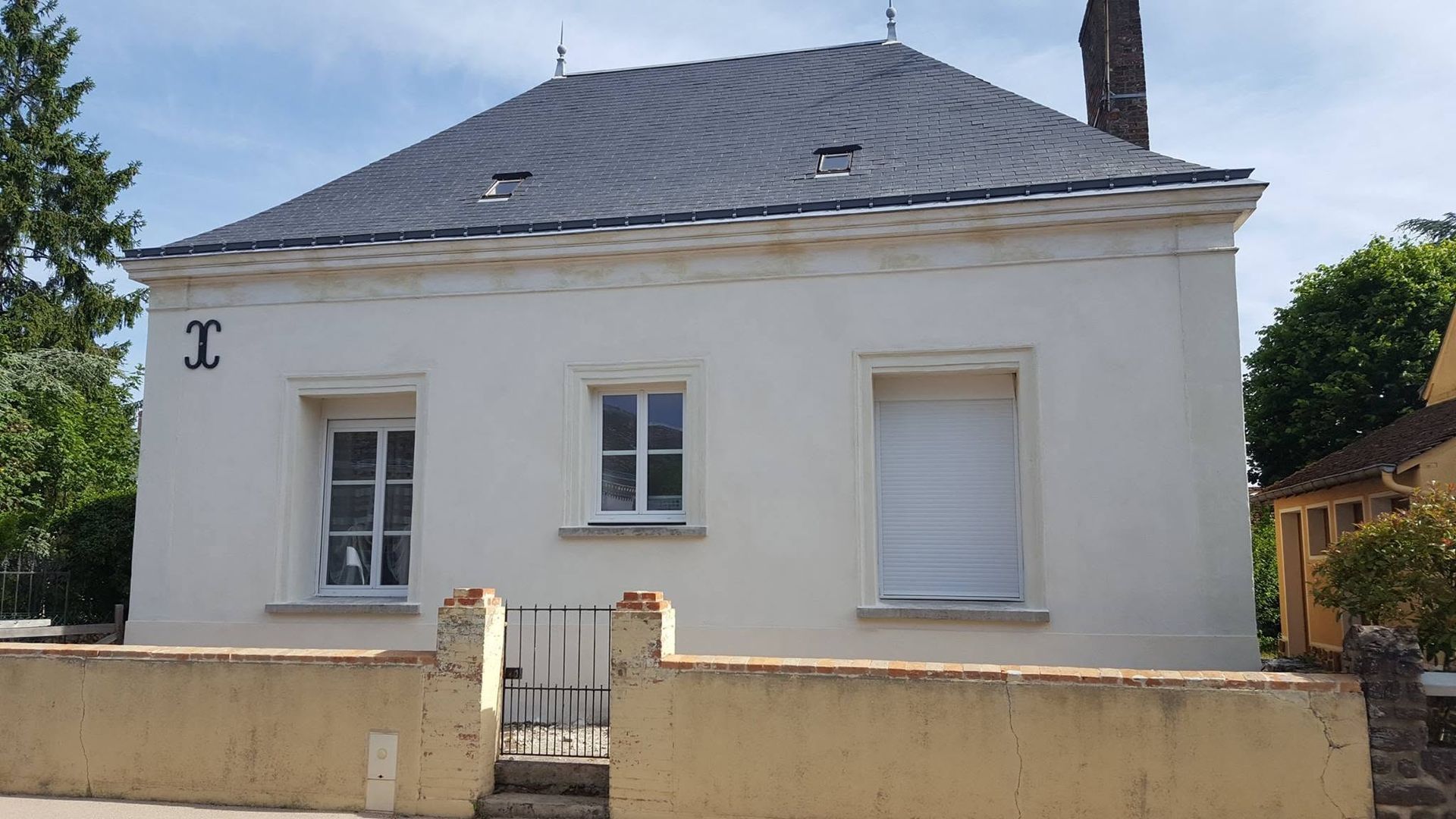Maison blanche avec un toit gris, deux fenêtres en façade et un muret beige bas devant.
