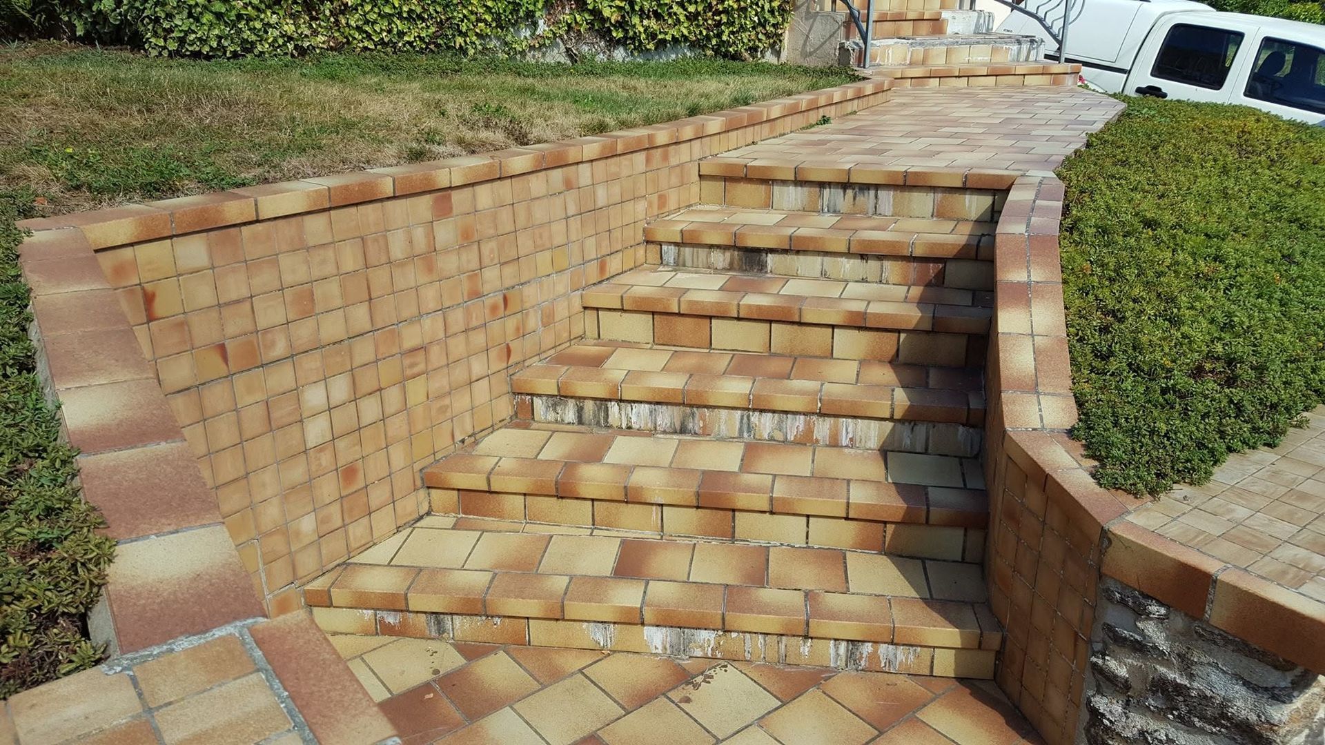 Escalier en briques intégré à un mur de soutènement paysager extérieur, avec des arbustes et un véhicule stationné en haut.