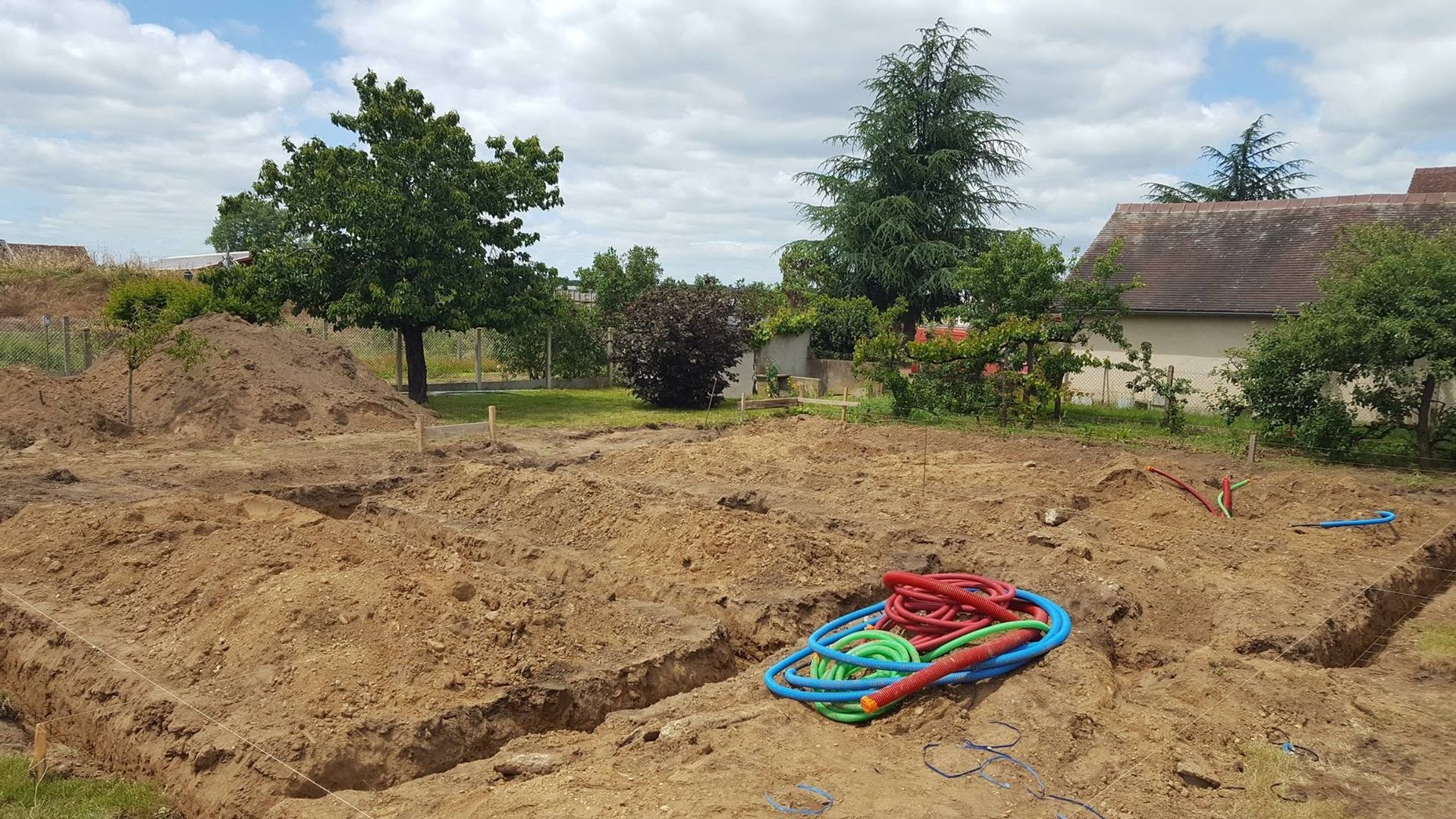 Des tas de sable dans une cour avec un jouet en plastique coloré et des arbres près d'une maison sous un ciel nuageux.