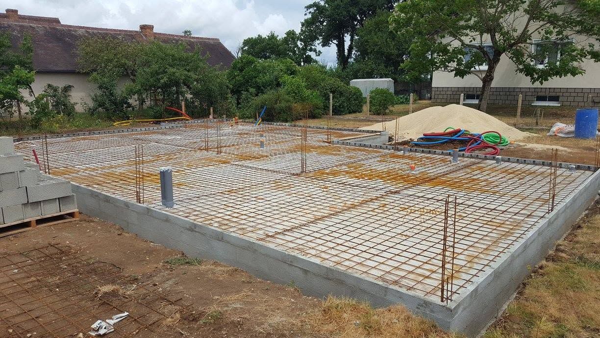 Fondation en béton en construction avec des rangées d'armatures et de sable dans un terrain extérieur clôturé.