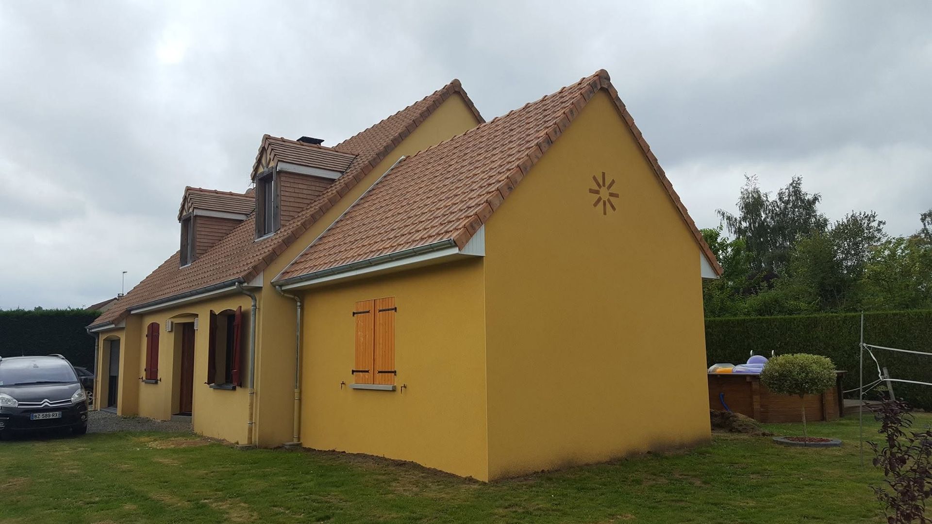 Maison jaune au toit de tuiles et aux fenêtres à volets, vue de côté sur un terrain herbeux sous un ciel nuageux.