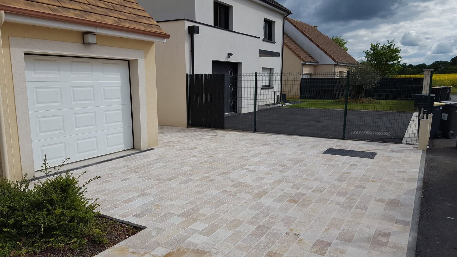 Allée pavée longeant un garage et une maison blancs, avec un portail et une clôture noirs devant.
