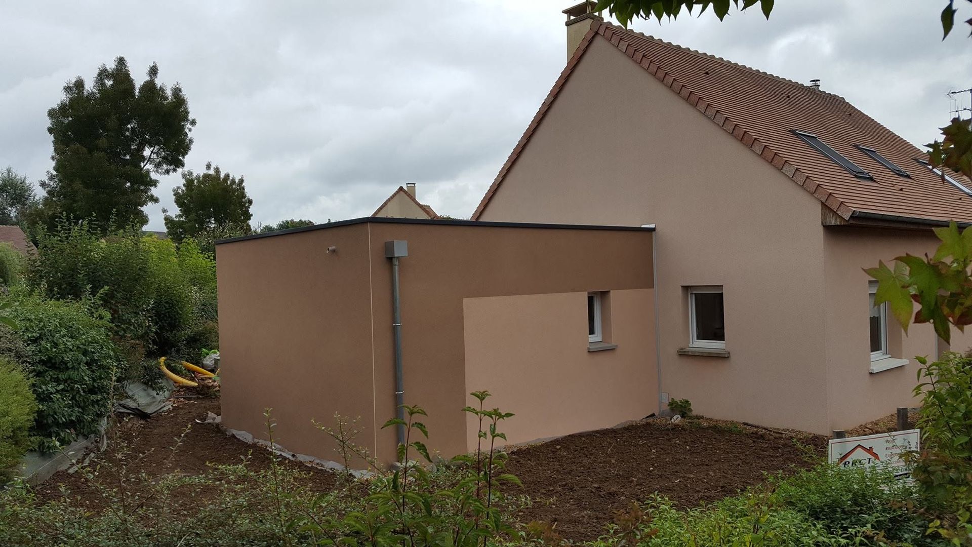 Petite maison beige au toit de tuiles rouges, jouxtant un jardin envahi par la végétation sous un ciel nuageux.