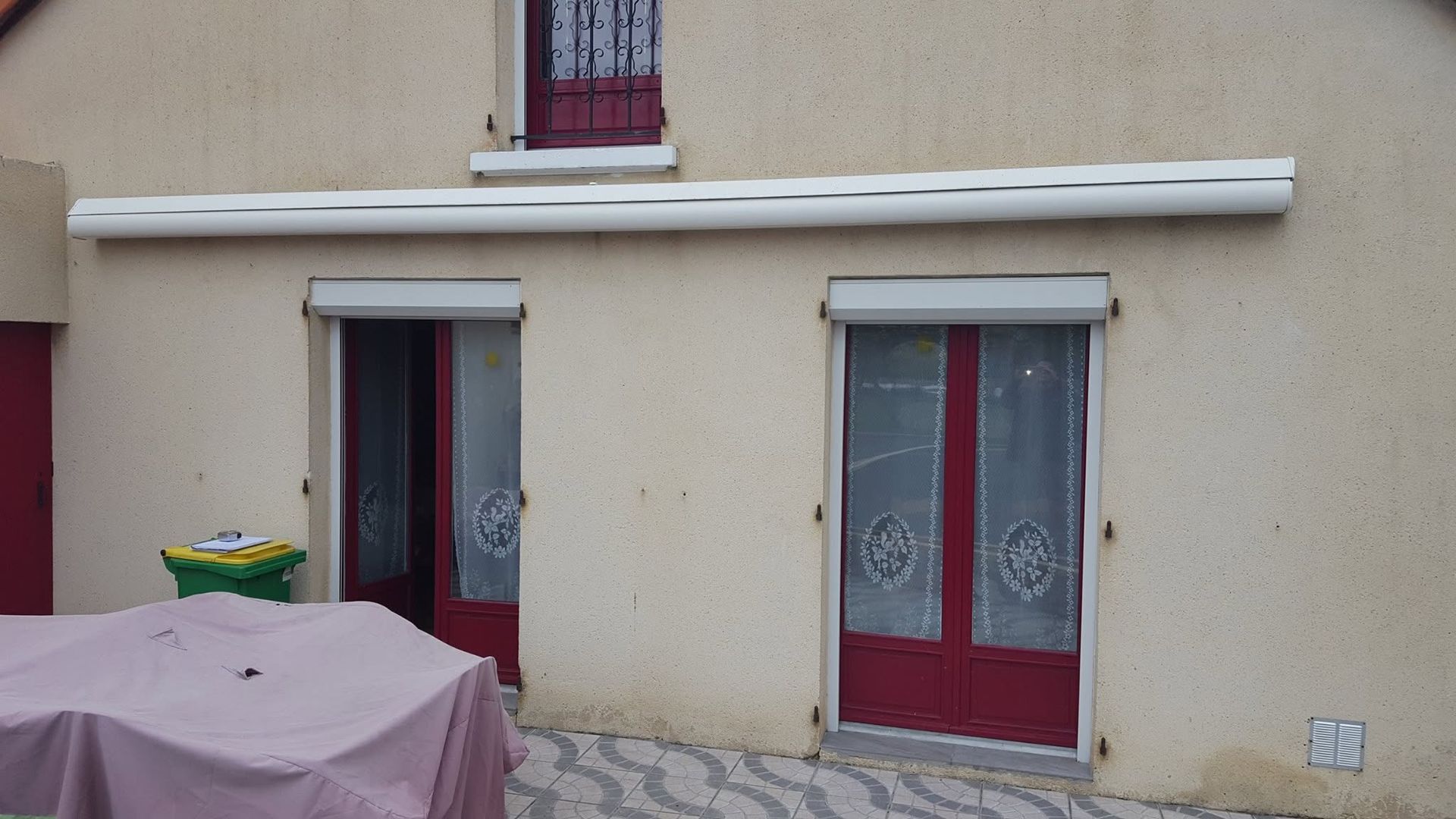 Façade beige de maison avec deux portes-fenêtres rouges, une petite fenêtre à l'étage et une table recouverte devant.