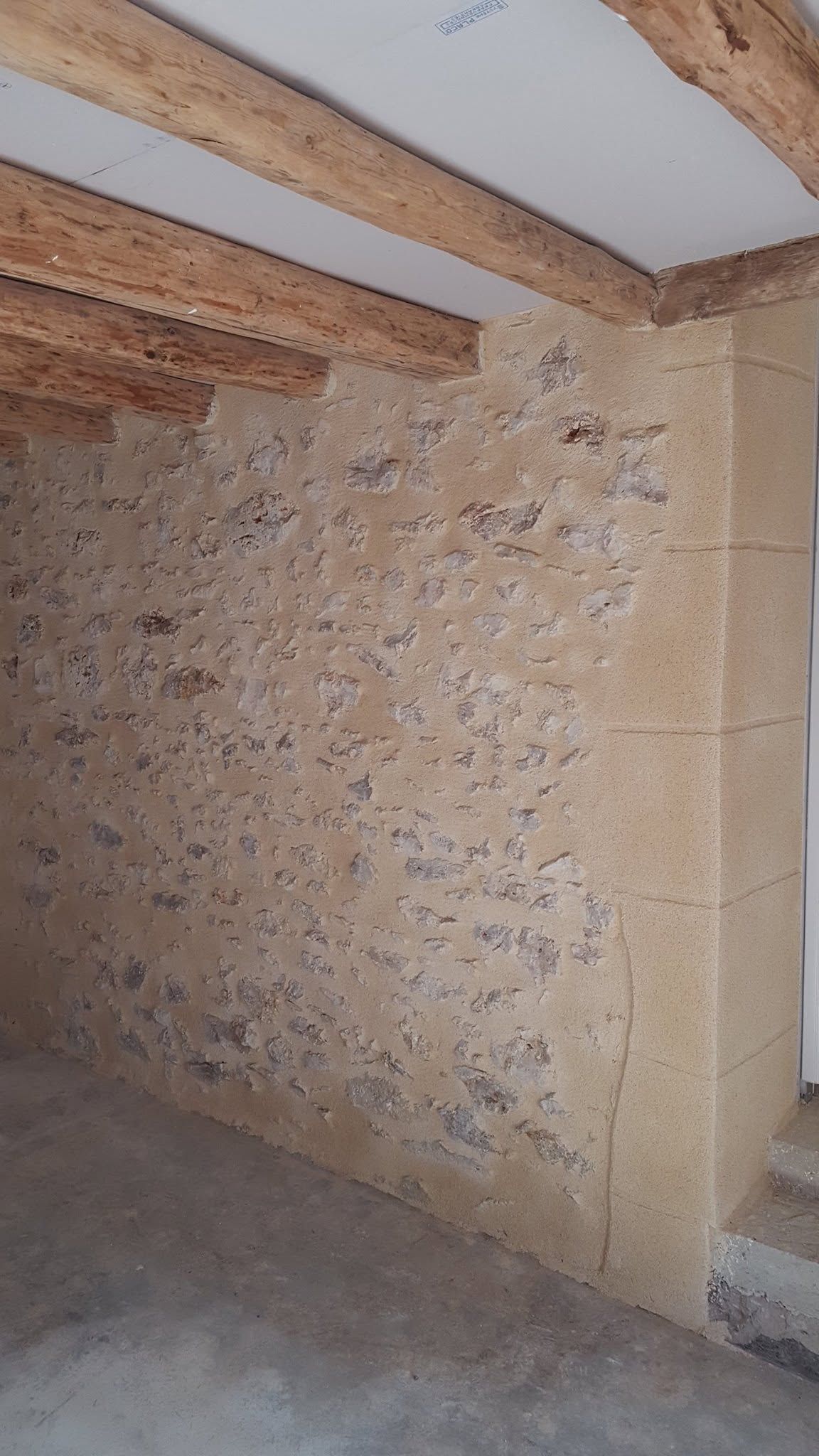 Pièce mansardée avec mur en pierre brute, poutres en bois apparentes et sol en béton brut.