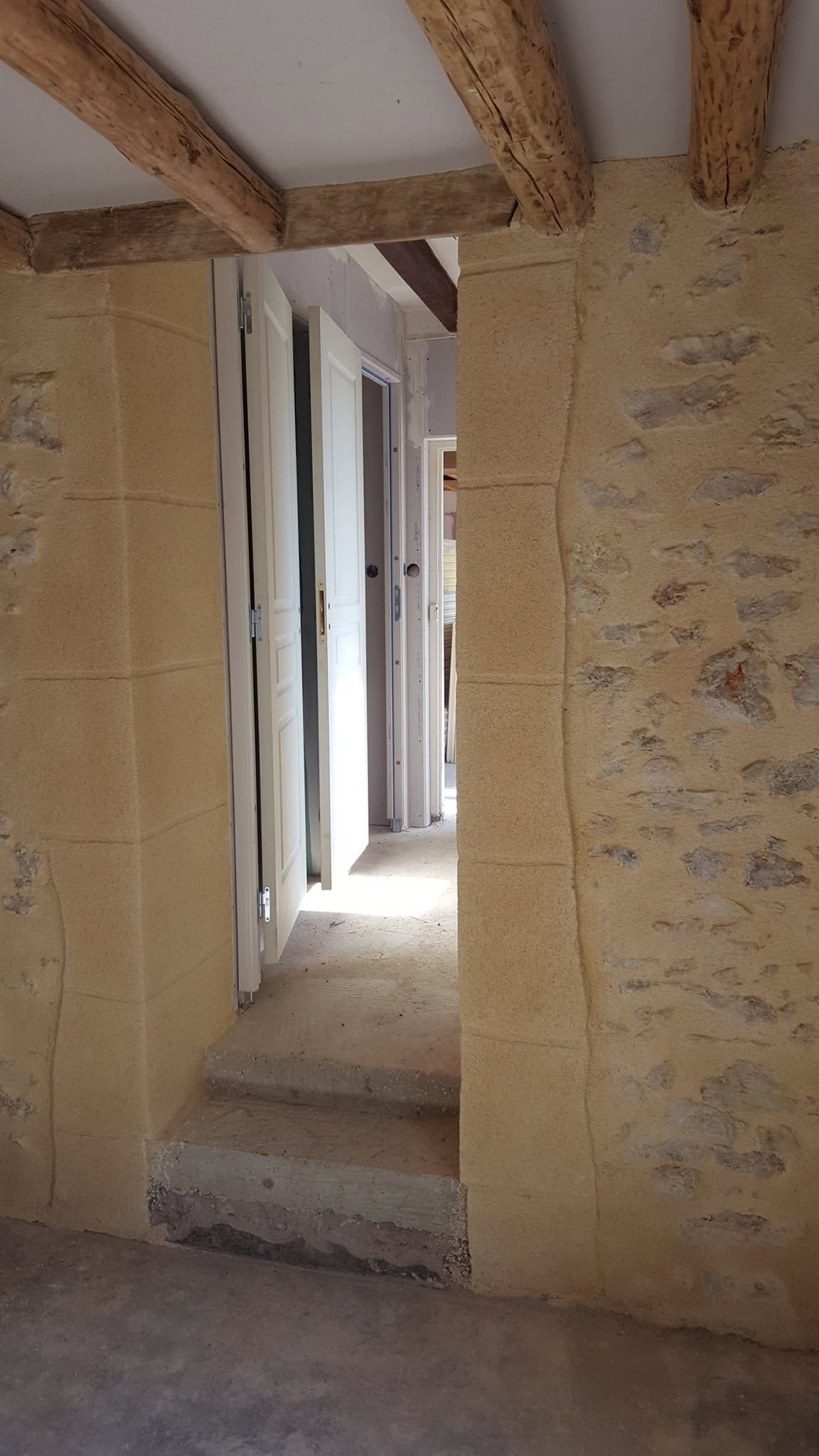 Couloir étroit aux murs de pierre, poutres en bois apparentes et un petit escalier menant à une porte lumineuse.