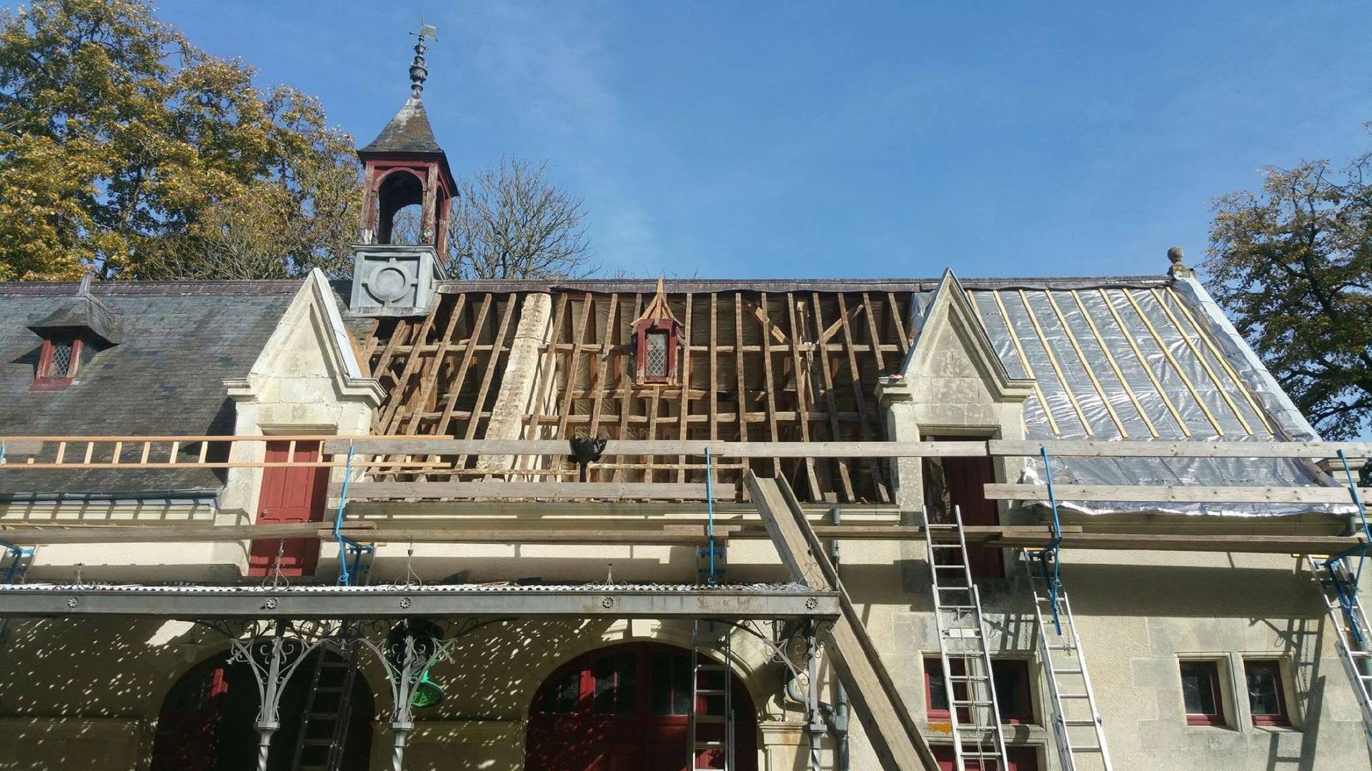Bâtiment historique en pierre en cours de rénovation, avec des échafaudages sur le toit et une petite tour d'horloge au sommet.