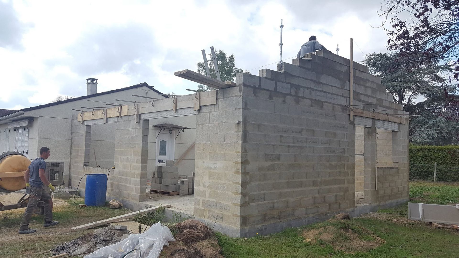 Maison en parpaings partiellement construite, avec des ouvriers et des matériaux de construction dans la cour.