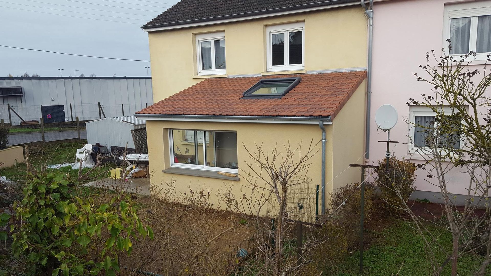 Maison jaune au toit de tuiles rouges et au jardin envahi par la végétation un jour de pluie.
