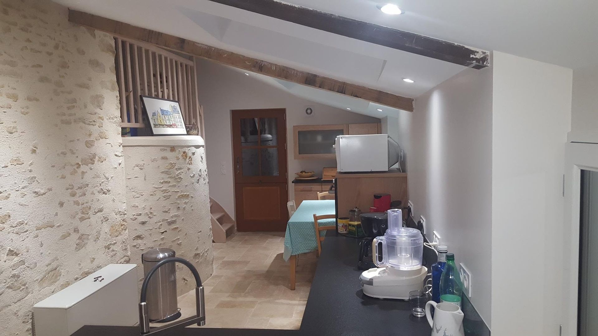 Petit espace cuisine-séjour avec plafond mansardé, comptoir foncé, sol carrelé et une table près du fond.