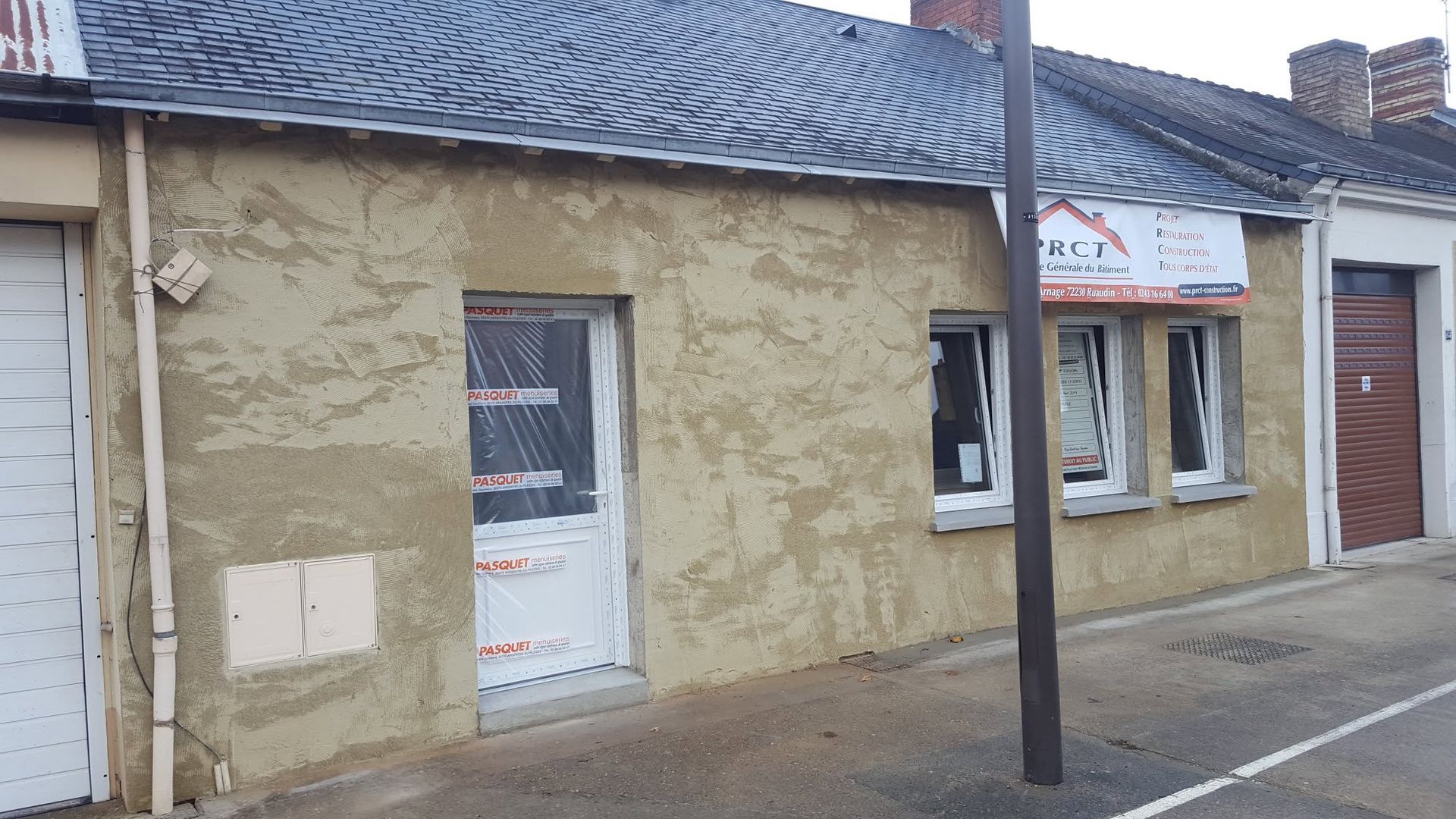 Une rangée de petites boutiques aux murs beiges défraîchis, aux portes et fenêtres blanches, et un lampadaire noir sur une rue mouillée.