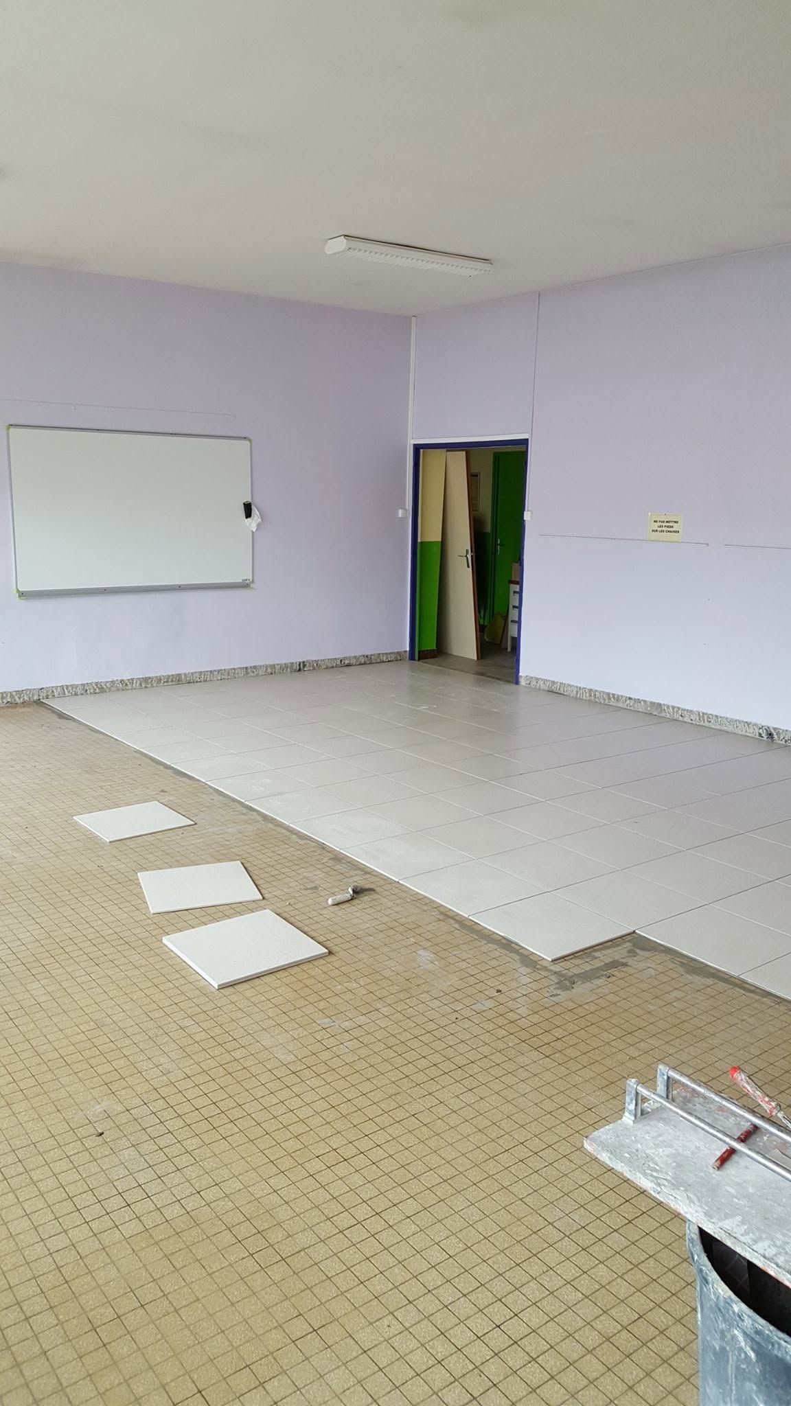 Salle de classe vide avec tableau blanc, murs lavande, sol carrelé et papiers étalés au sol.