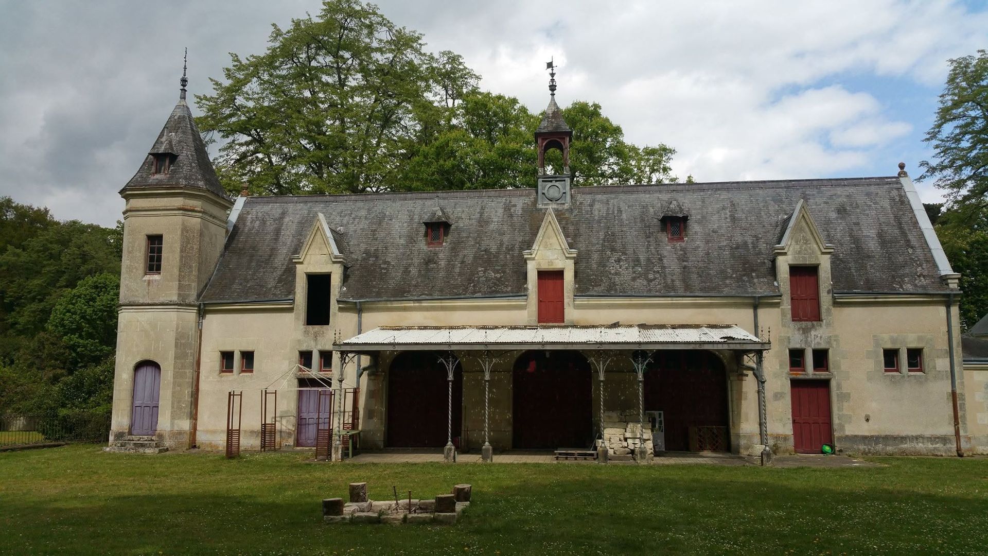 Vieille demeure beige avec une tour et une longue véranda, située dans une cour herbeuse sous un ciel nuageux.