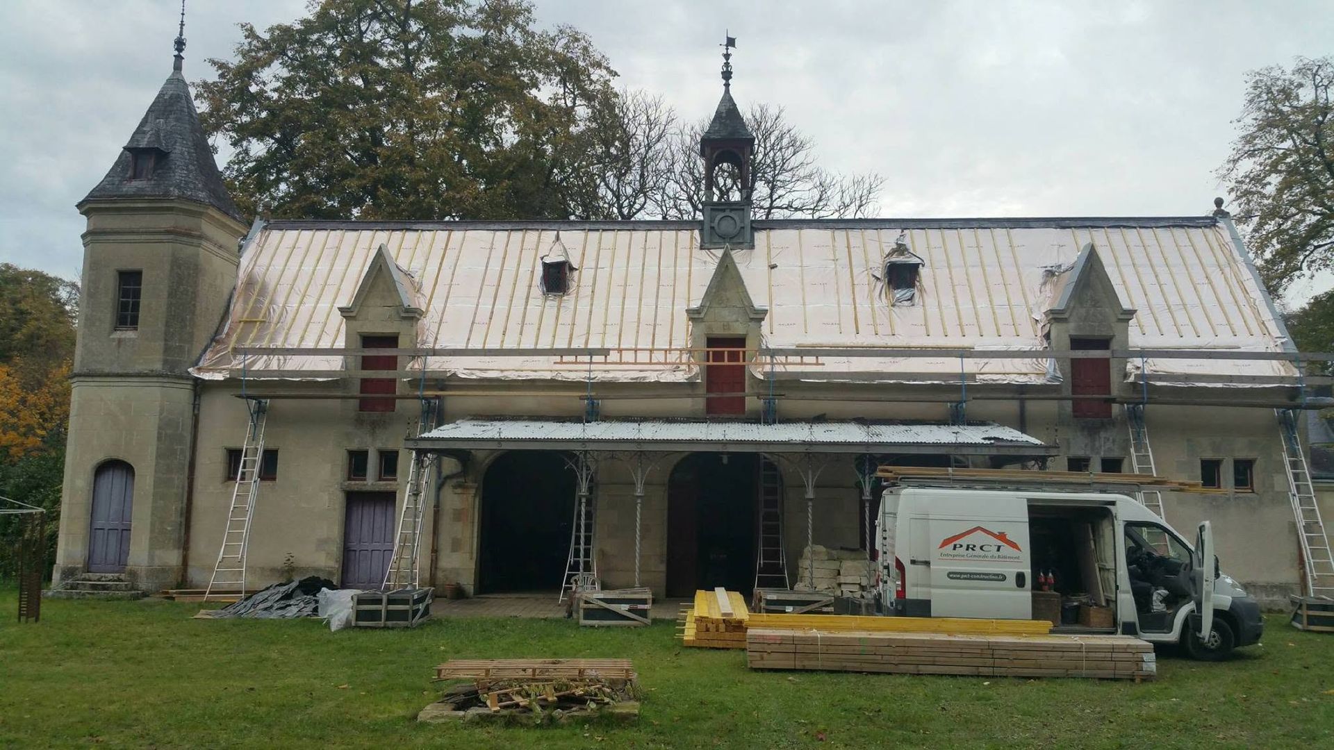 Bâtiment historique en pierre avec toit orné, entrée principale ouverte et camion garé à l'extérieur sur une pelouse.