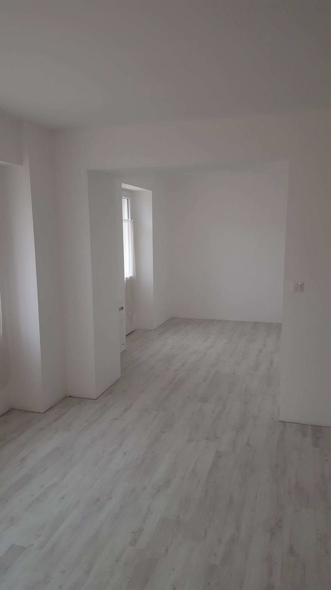 Pièce blanche vide avec parquet clair et une porte donnant sur une autre pièce.