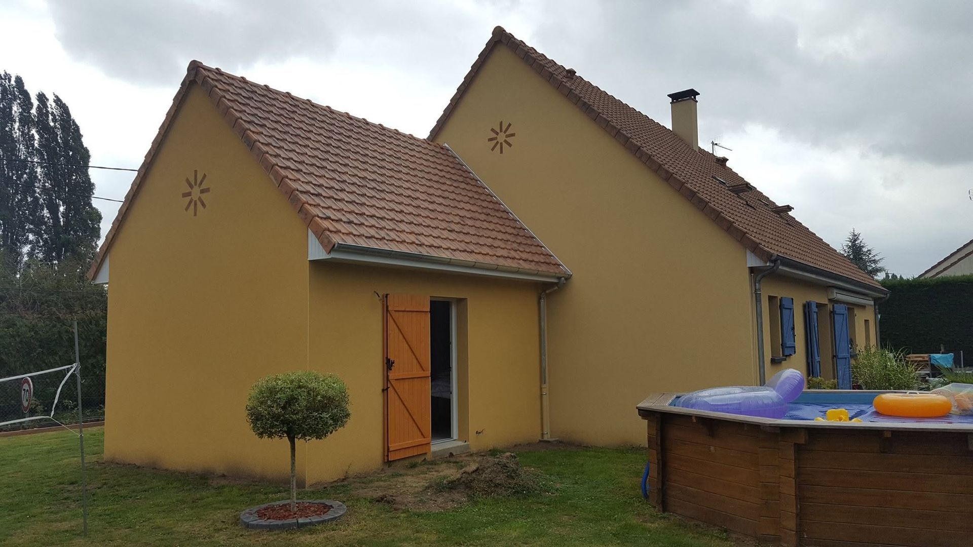 Maison jaune au toit de tuiles rouges à côté d'une petite piscine hors sol sous un ciel nuageux.