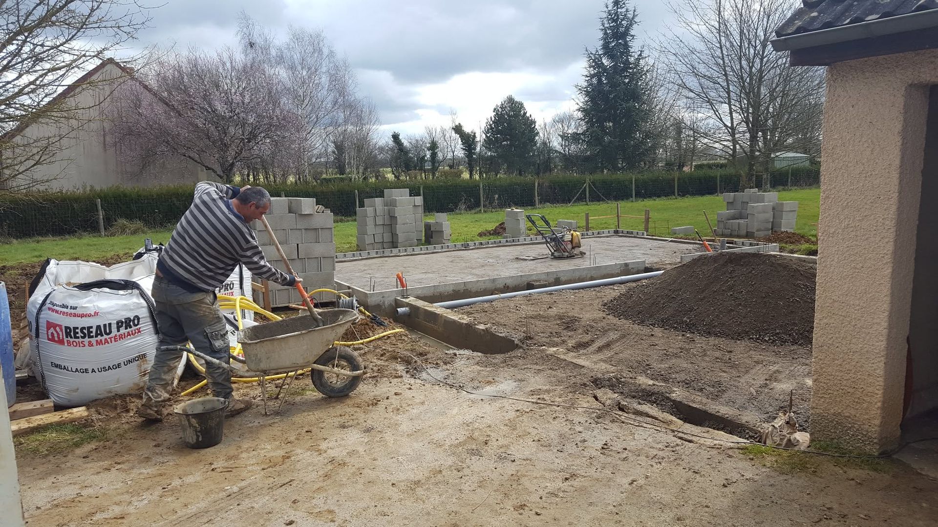Un ouvrier manœuvre une petite excavatrice à côté d'une maison, creusant une tranchée dans une cour boueuse.