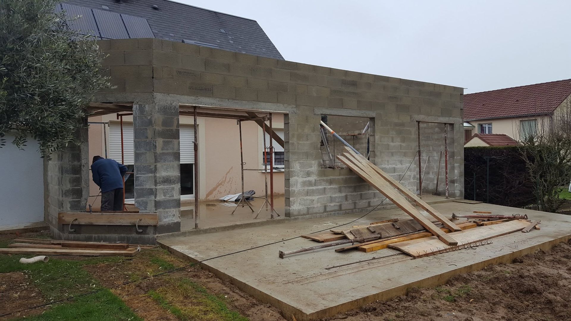 Maison en construction avec des murs en béton inachevés, des supports en bois et un ouvrier sur la gauche.