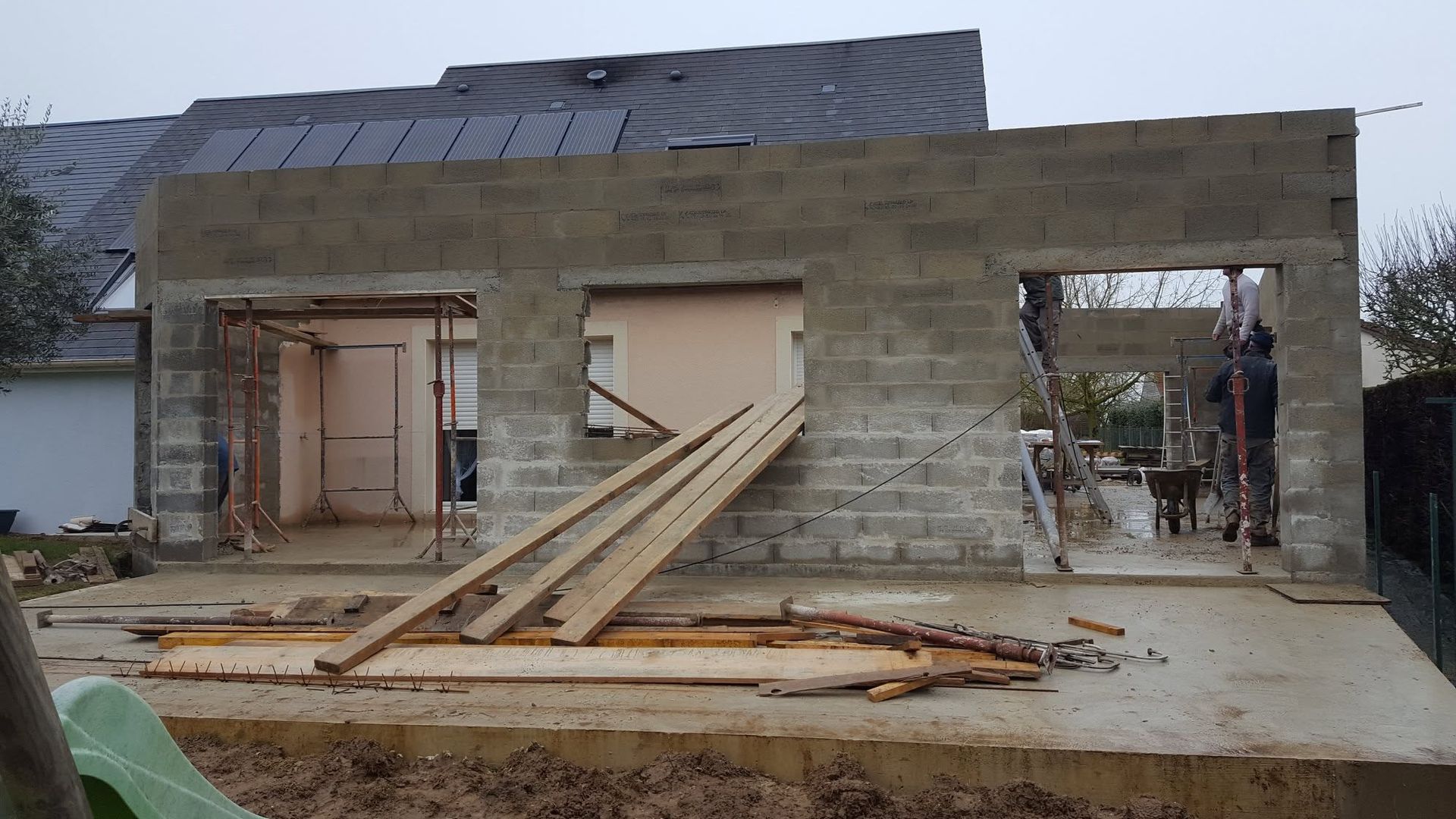 Maison en béton partiellement construite, avec portes ouvertes, toiture en construction et planches de bois empilées devant.