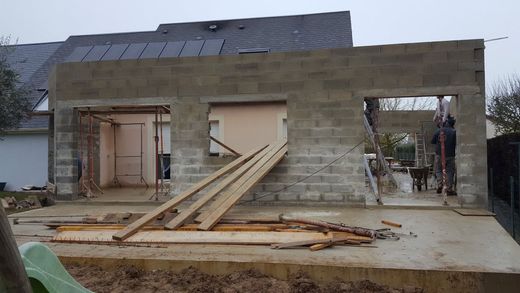 Maison en béton inachevée en construction, avec des portes ouvertes et des planches de bois appuyées contre le mur de façade.