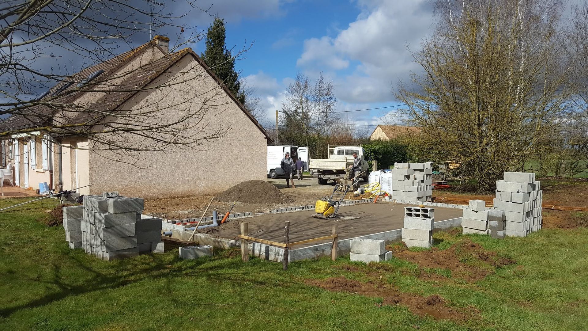 Maison en pierre partiellement construite, avec des ouvriers et des blocs empilés dans une cour herbeuse sous un ciel nuageux.