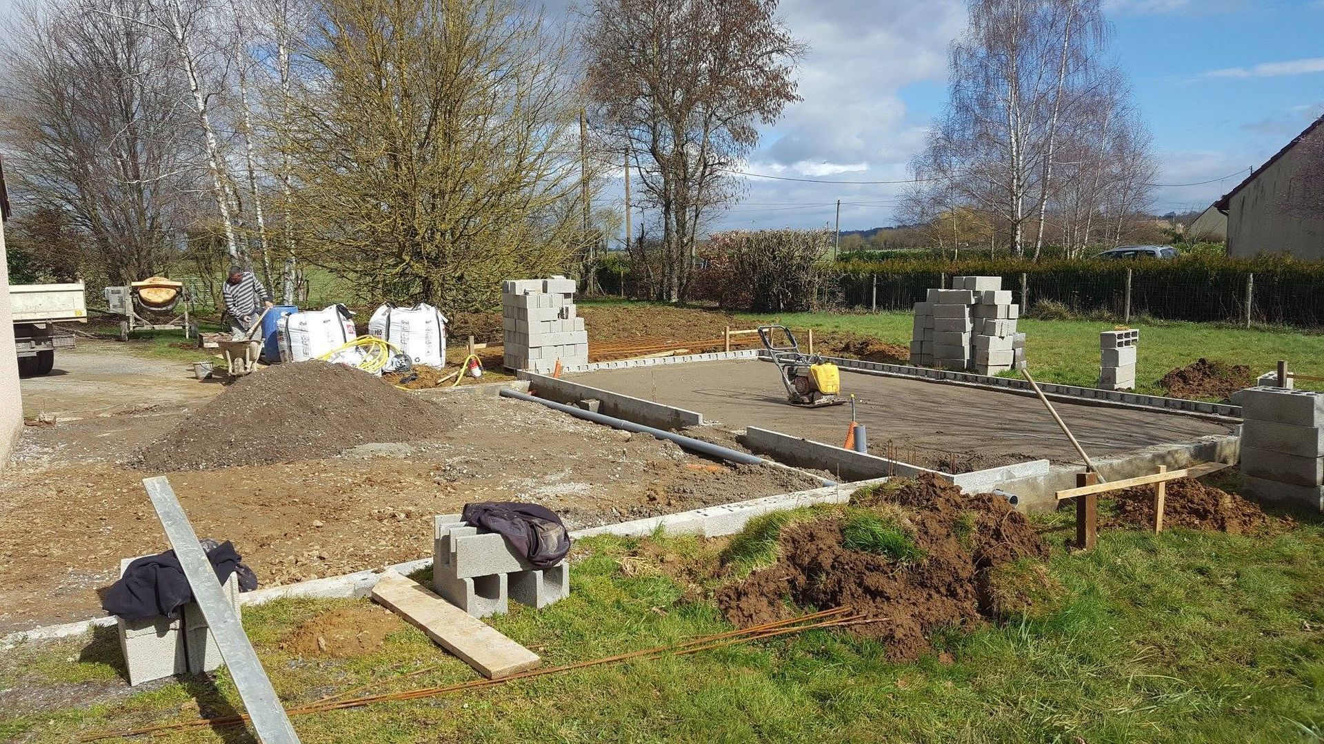 Fondations en béton partiellement construites dans une cour rurale, avec des outils et des matériaux sous un ciel ensoleillé.