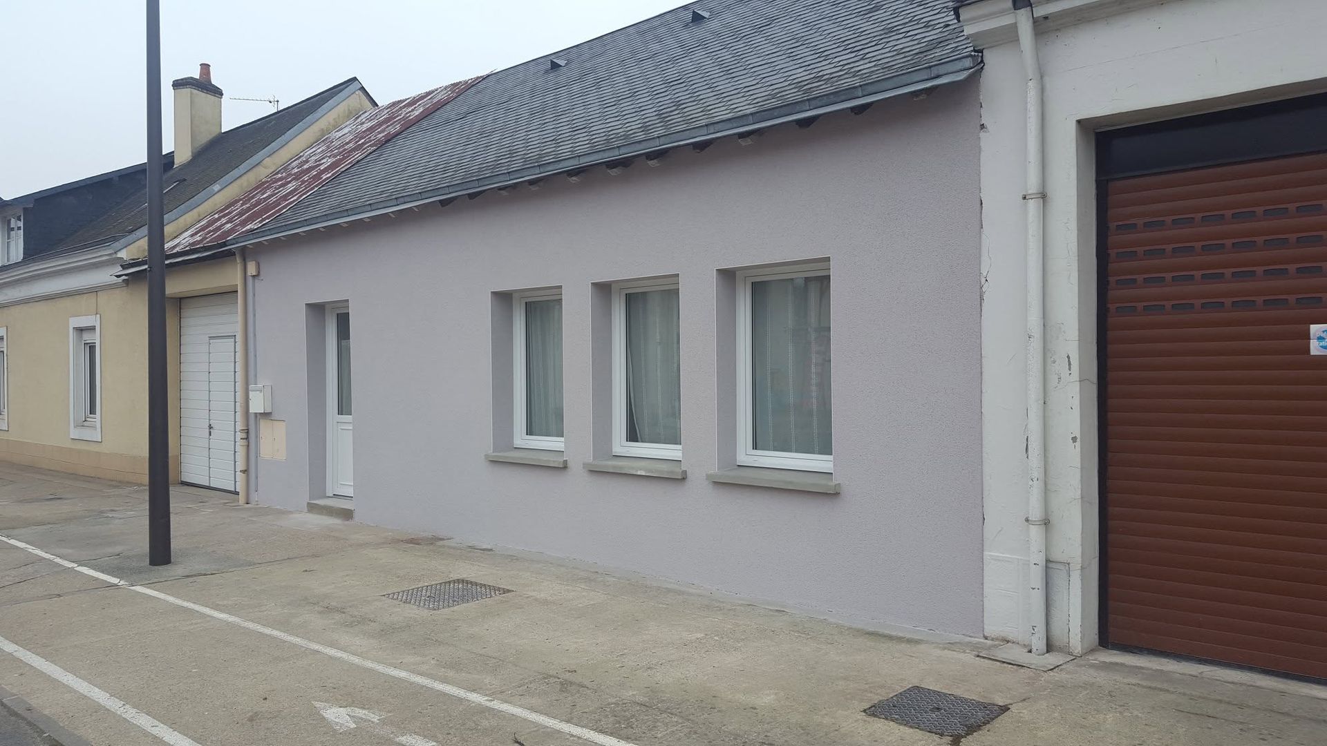 Façade grise d'une maison avec trois fenêtres, un lampadaire et une grande porte de garage, donnant sur une rue calme.