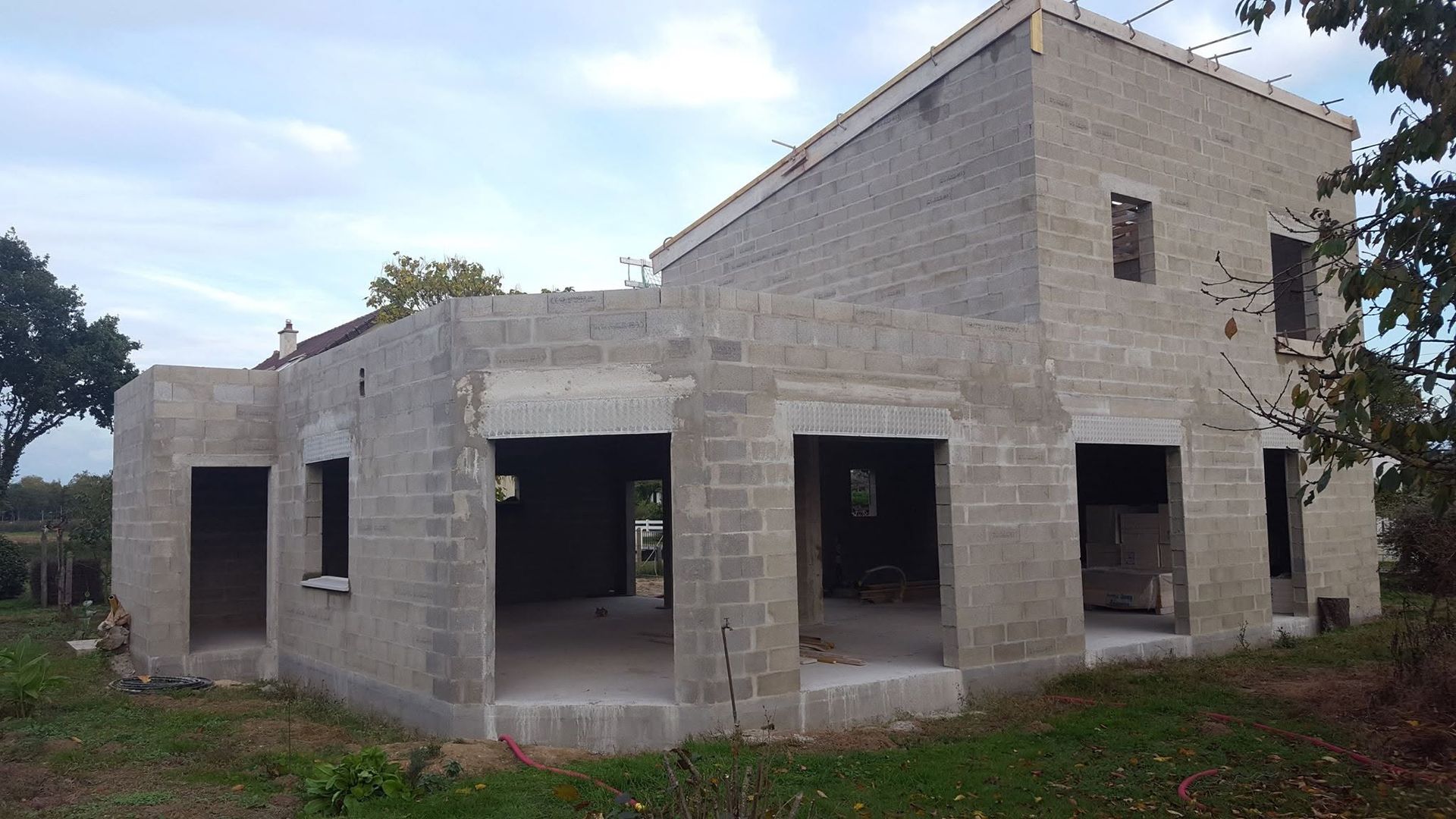 Maison inachevée en béton gris, avec portes et fenêtres ouvertes, dans une zone rurale herbeuse sous un ciel nuageux.
