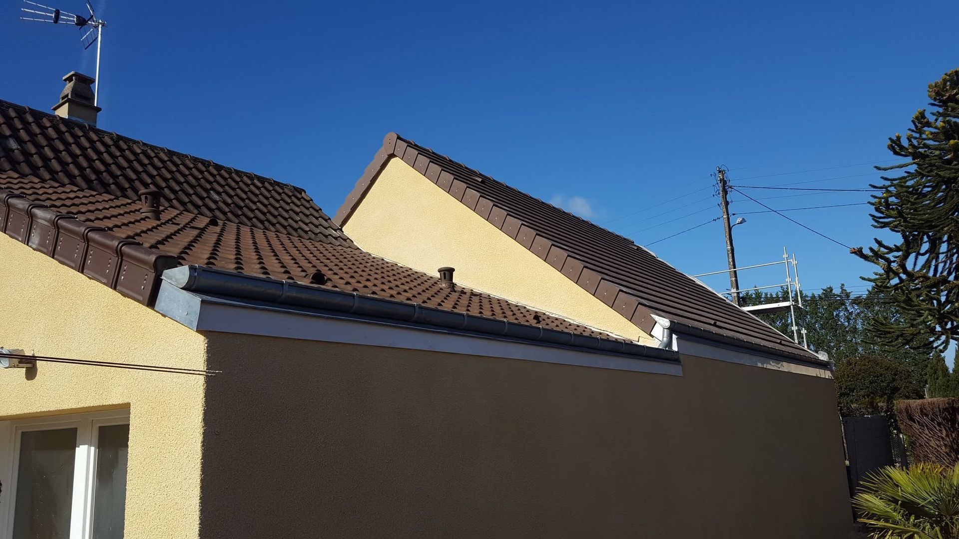 Toits de banlieue et murs beiges sous un ciel bleu clair, avec une antenne de télévision et des arbres en arrière-plan.
